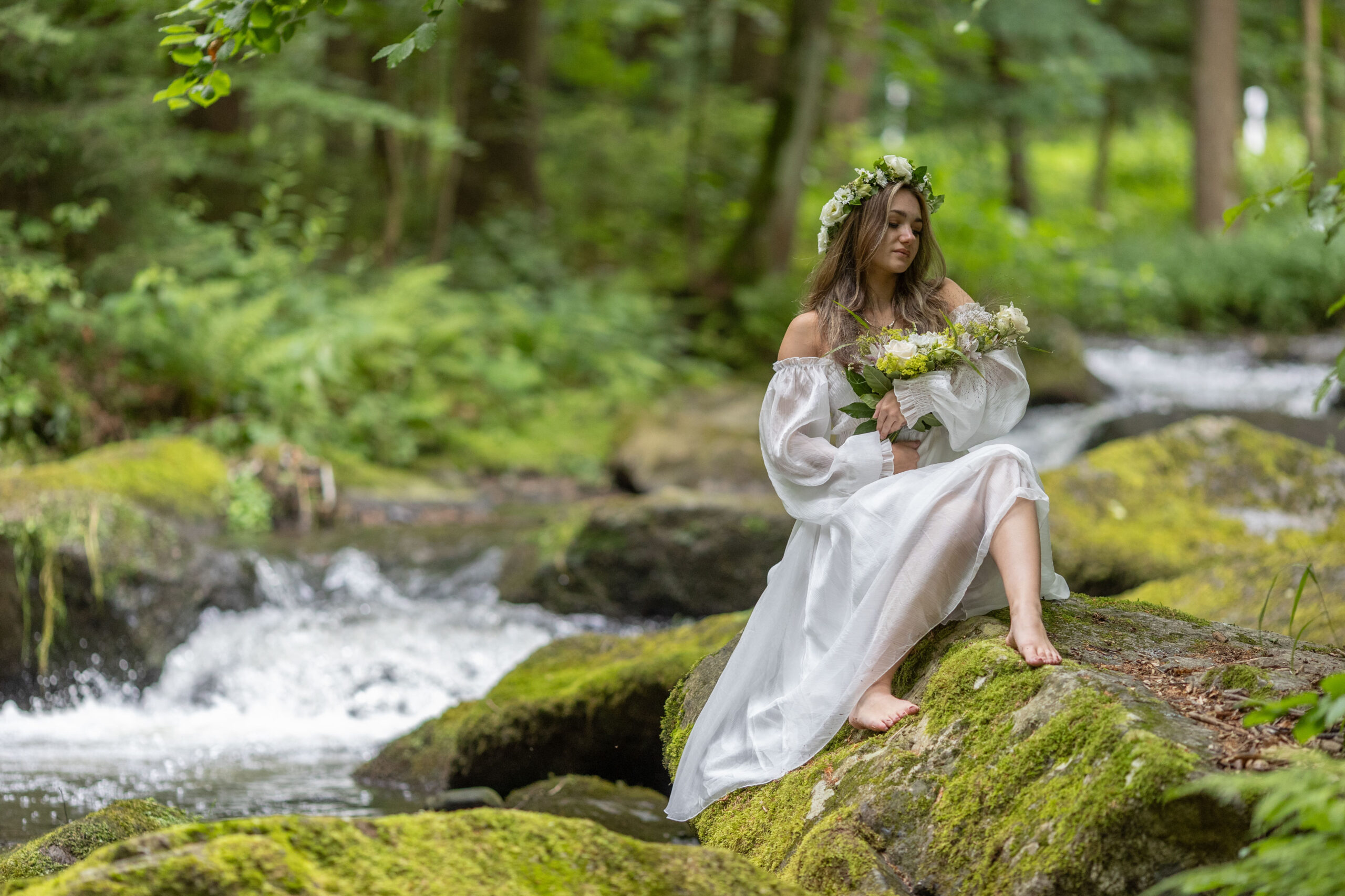 Porträt einer jungen Frau in weißem Kleid mit Blumenkranz im Haar sitzt auf einem Stein eines Baches im Erzgebirge