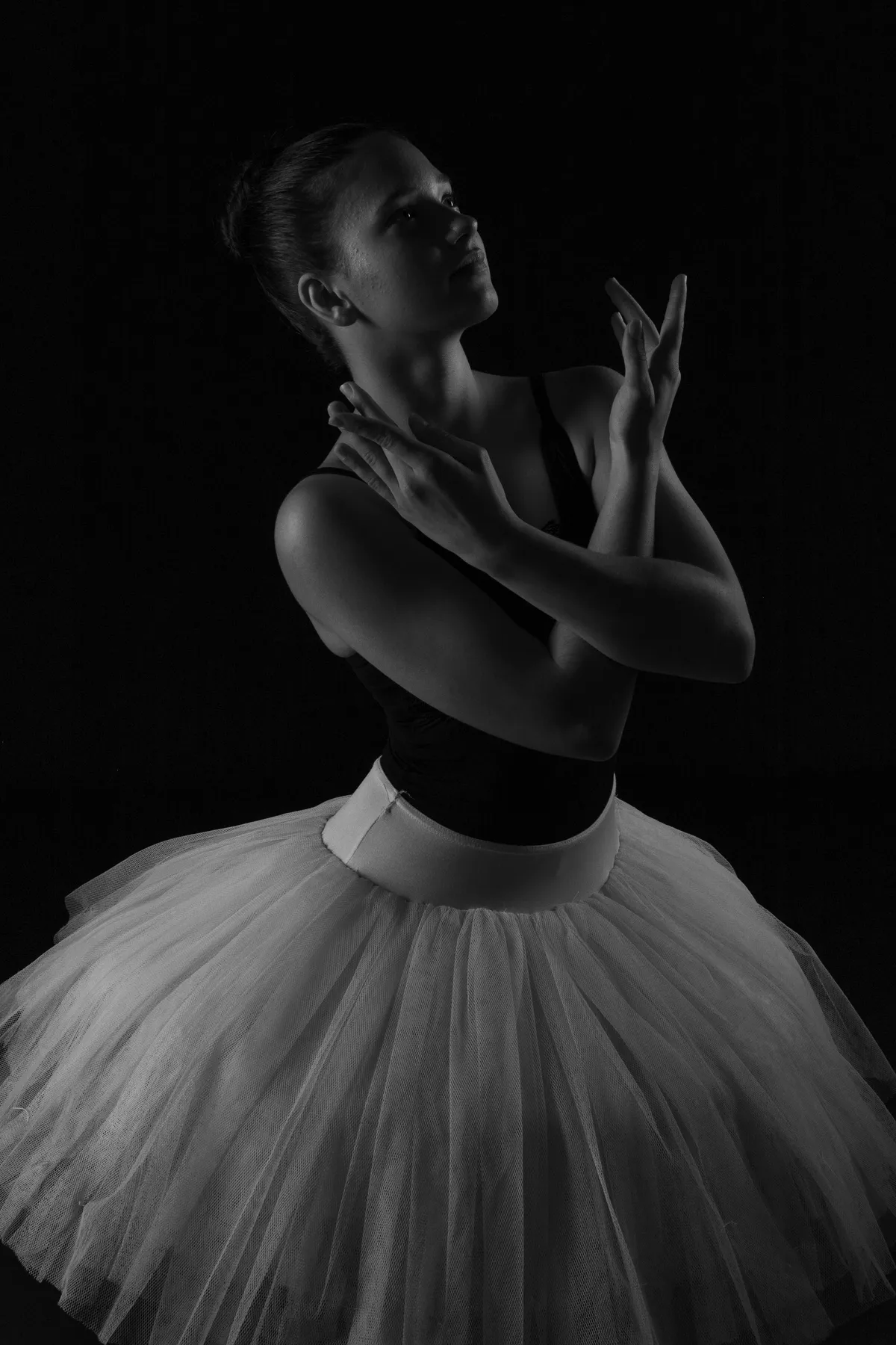 Eine Ballerina in einem weißen Tutu posiert anmutig mit gekreuzten und erhobenen Armen und blickt nach oben vor einem dunklen Hintergrund. Die Beleuchtung hebt ihr Gesicht und ihr Kostüm hervor und schafft eine dramatische, elegante Silhouette.