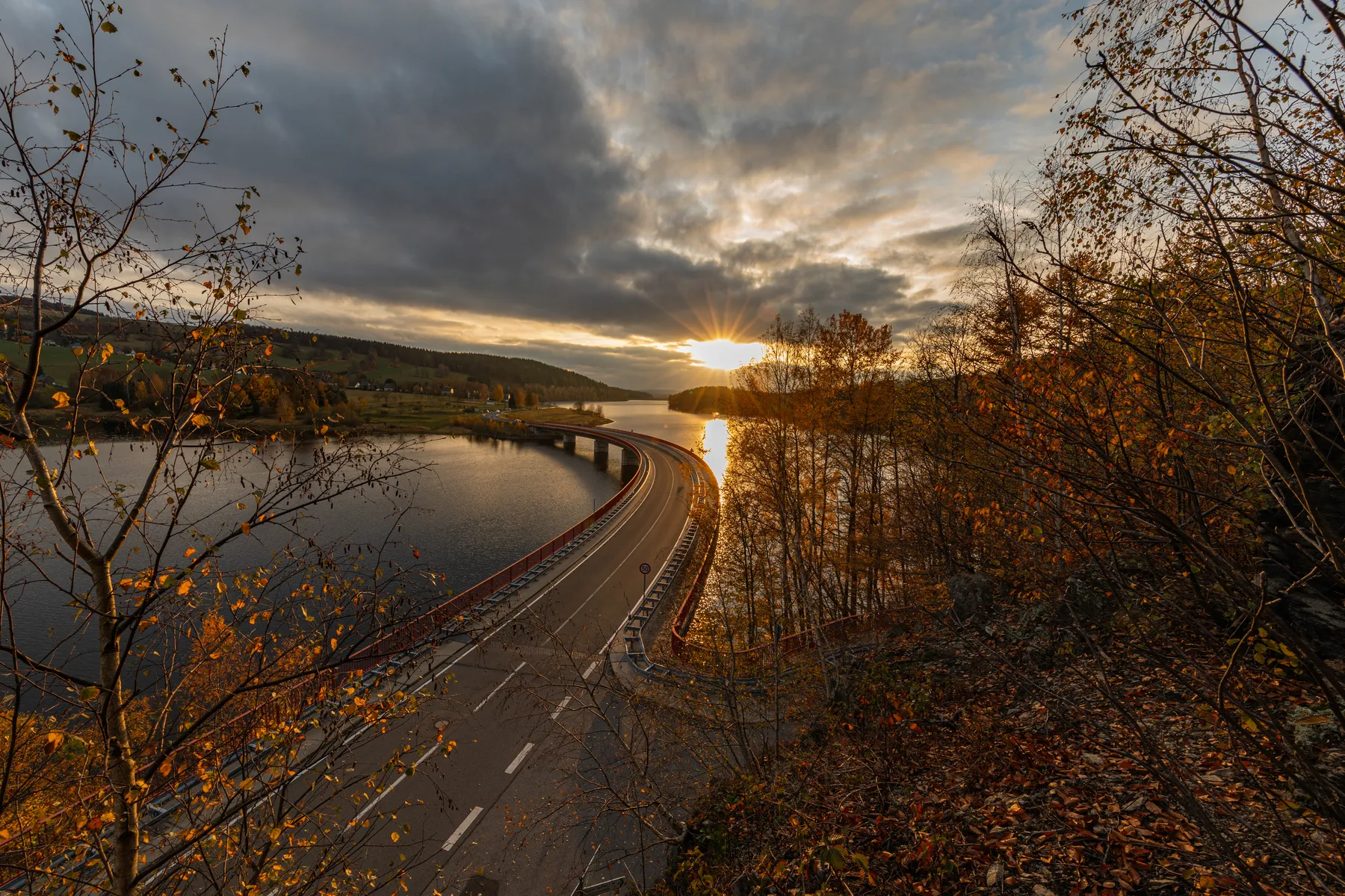Eine kurvenreiche Straße führt über die Rauschenbachtalsperre im Erzgebirge, wobei die Sonne teilweise von Wolken verdeckt wird. Bäume mit Herbstlaub rahmen die Szene ein und reflektieren das goldene Licht auf dem Wasser welches ruhig ist.
