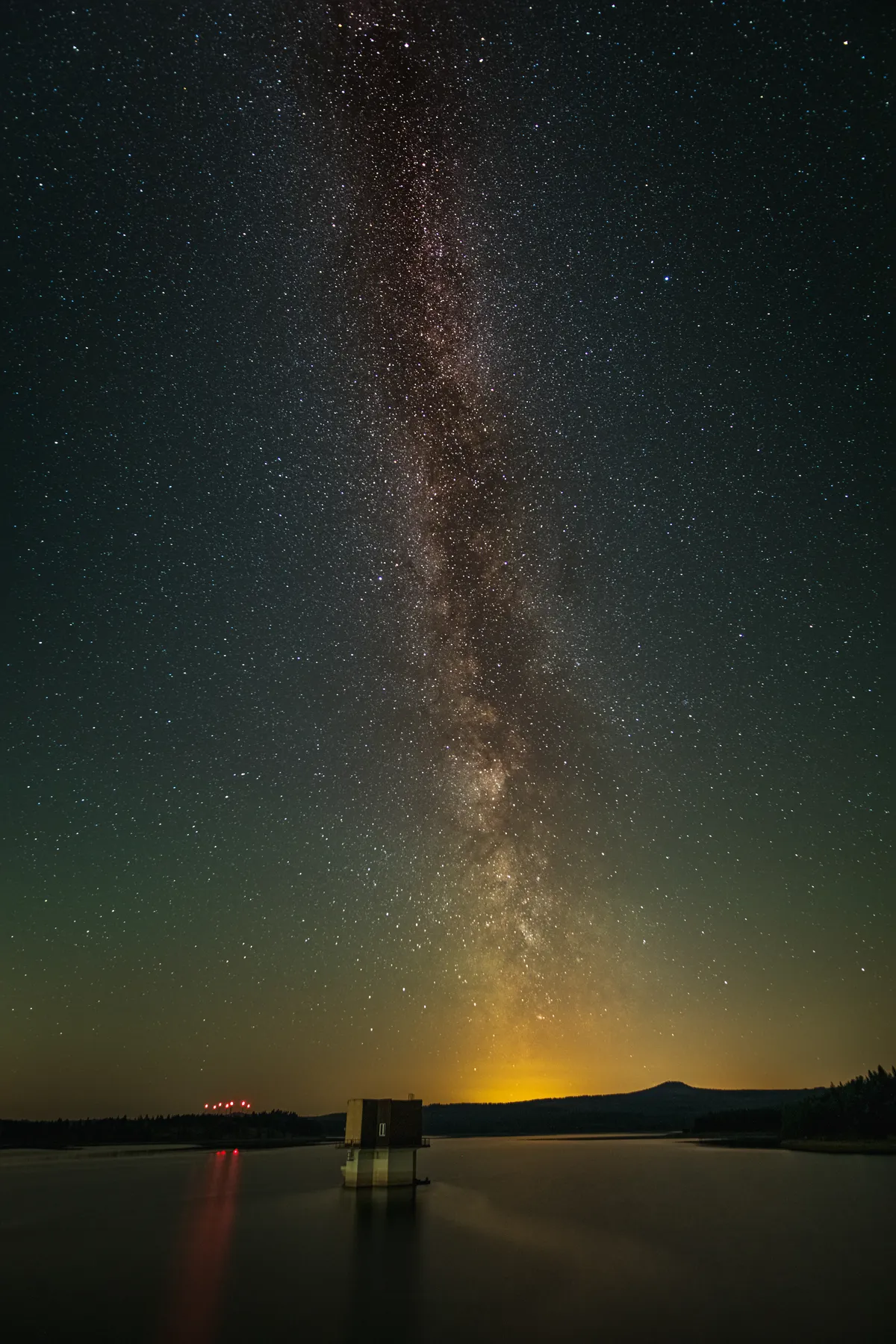 Der Nachthimmel über der Preßnitztalsperre im Böhmischen Erzgebirge leuchtet mit Sternen und der Milchstraße, die sich auf dem ruhigen Wasser spiegelt. Eine kleine dunkle Struktur, die auf bewaldeten Hügeln schwimmt, und ein schwach orangefarbener Horizont bilden die perfekte Kulisse für dieses Foto