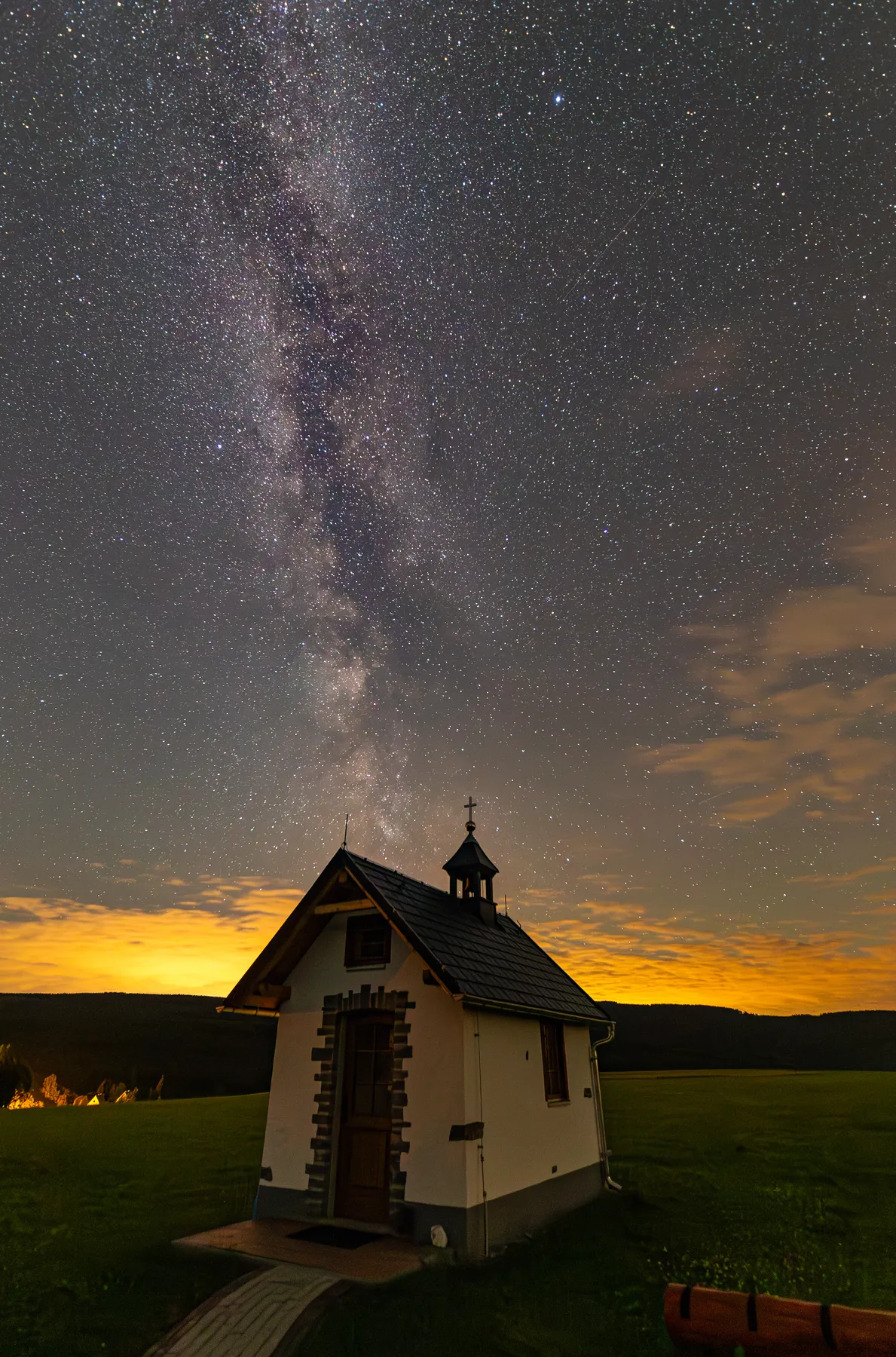 Die Wandererkapelle von Kretscham-Rothensehma steht auf einer grasbewachsenen Wiese unter der leuchtenden Milchstraße. Am Horizont leuchten warme Lichter, während sich dunkle Hügel als Silhouetten unter dem sternenklaren Nachthimmel abzeichnen.