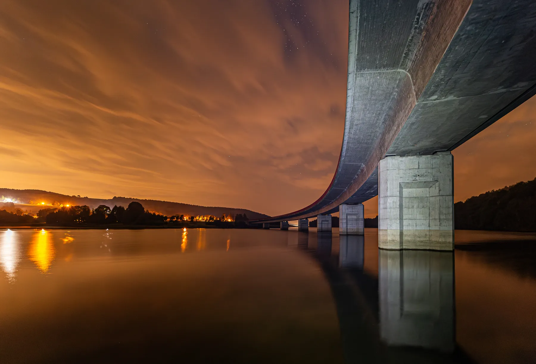 Eine große Betonbrücke führt durch die Rauschenbachtalsperre im Erzgebirge, das ruhige Wasser in der Nacht, beleuchtet von orangefarbenen Lichtern und Spiegelungen; der Himmel ist wolkenverhangen mit einem warmen Schein, und Bäume und einzelne Häuser sowie Laternen säumen den Horizont.