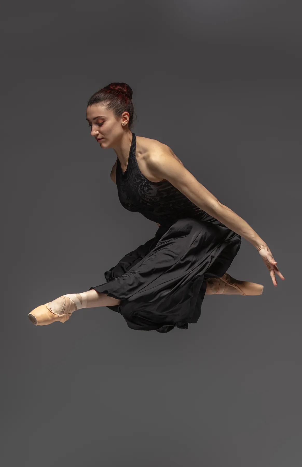 Eine Balletttänzerin in einem schwarzen Kleid vollführt einen Sprung in der Luft mit spitzen Zehen und anmutig ausgestreckten Armen, eingefangen von einem erfahrenen Ballettfotografen vor einem schlichten grauen Hintergrund.