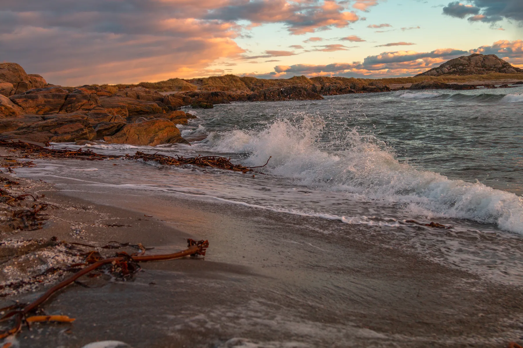 Bei Sonnenuntergang und teilweise bewölktem Himmel prallen die Wellen an der Südküste Norwegens auf eine felsige Küste. Seetang liegt auf dem nassen Sand verstreut, und goldenes Licht beleuchtet die Felsen und Wolken in der Ferne.