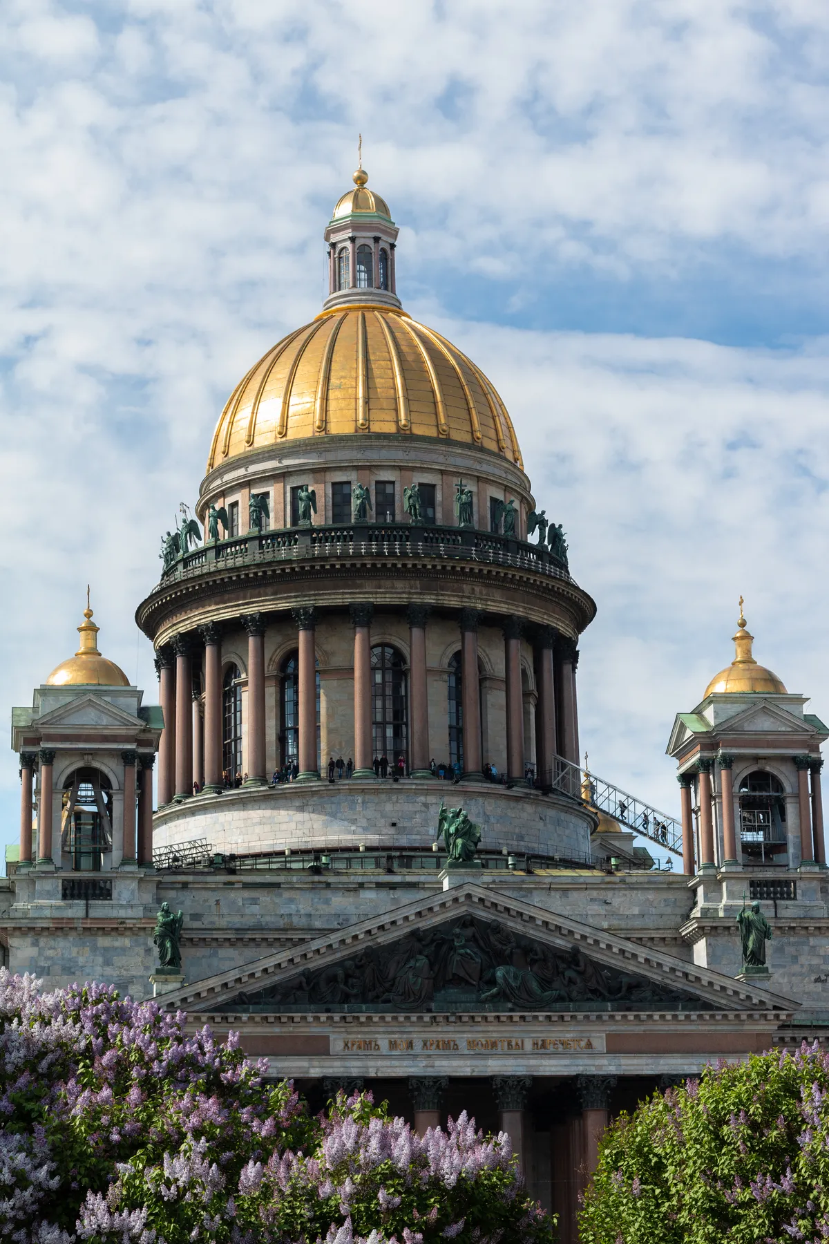 Nahaufnahme der mit einer goldenen Kuppel versehenen St. Isaakskathedrale in St. Petersburg, Russland, mit verschnörkelten Statuen auf dem Dach, gemeißelten Reliefs und einer Inschrift in russischer Sprache über dem Eingang, mit einem blauen Himmel im Hintergrund.