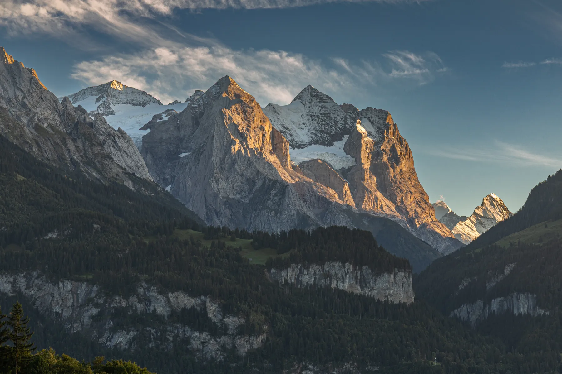 Majestätische Berggipfel mit Schneekappen, die vom warmen Sonnenlicht angestrahlt werden, erheben sich über dichte grüne Wälder und felsige Klippen unter einem blauen, wolkenverhangenen Himmel.