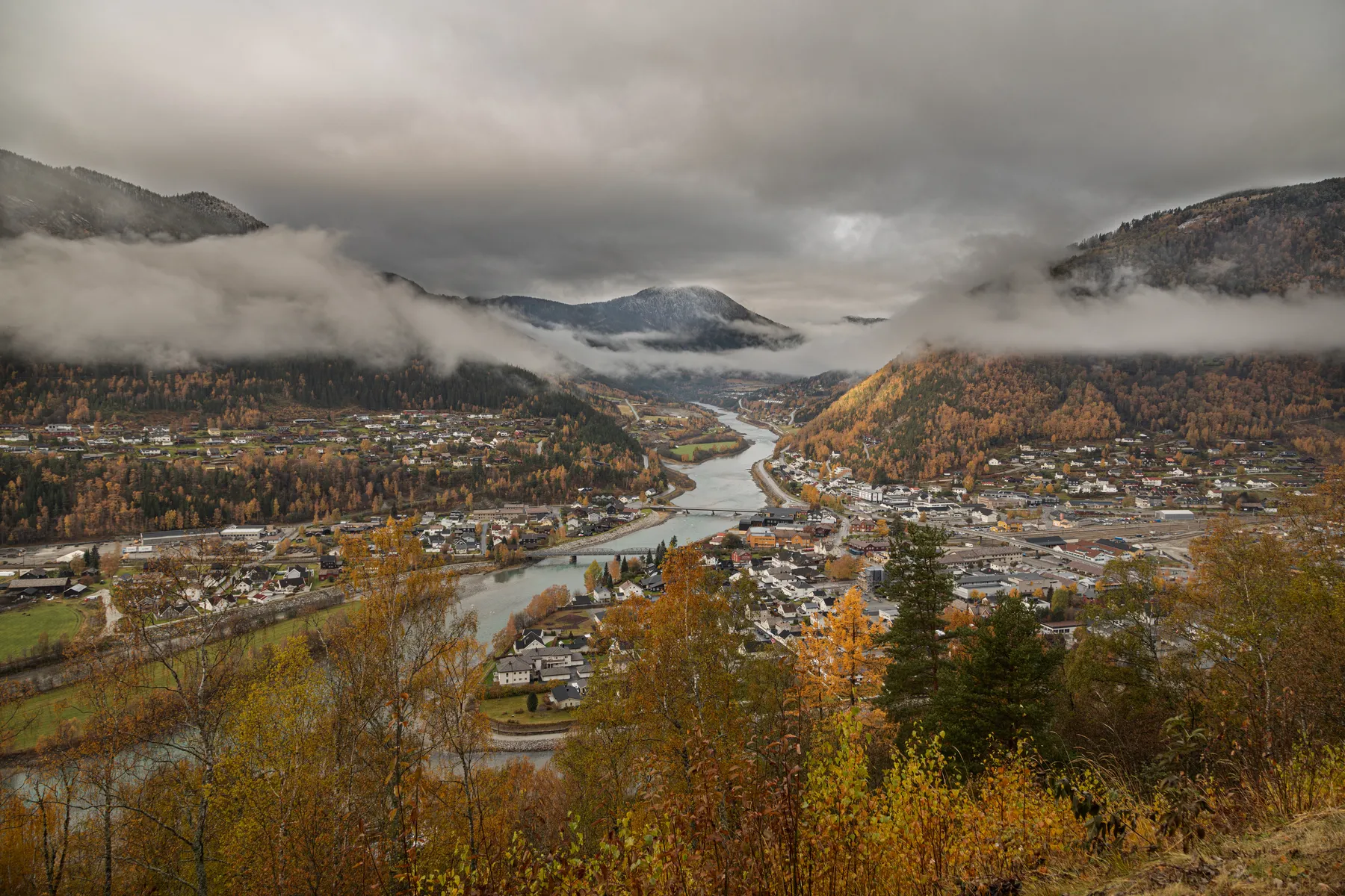 Ein Panoramablick auf eine kleine Stadt in Norwegen in einem Flusstal, umgeben von herbstlich gefärbten Bäumen und nebligen Bergen unter einem bewölkten Himmel.