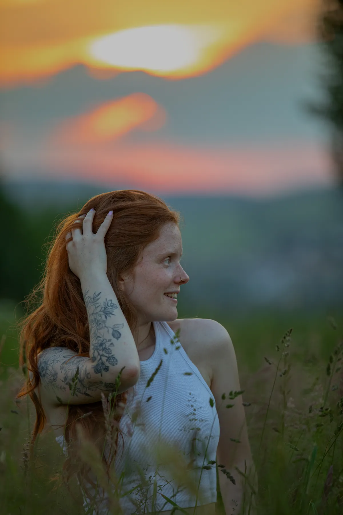Portraitfotografie einer jungen Frau mit langen roten Haaren und einer Blumentätowierung auf dem Arm bei Sonnenuntergang im hohen Gras. l