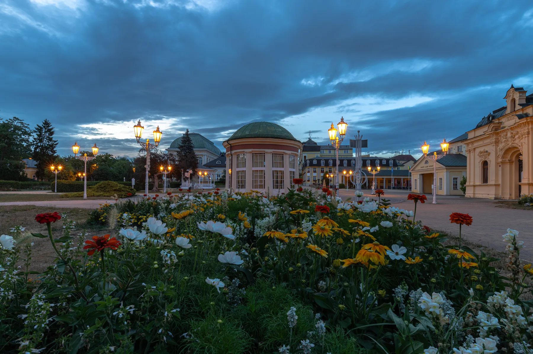 Ein Blumengarten mit roten, weißen und gelben Blüten befindet sich vor einem runden Gebäude mit einer grünen Kuppel. Verschnörkelte Straßenlaternen und historische Gebäude umgeben die Gegend unter einem bewölkten Abendhimmel.