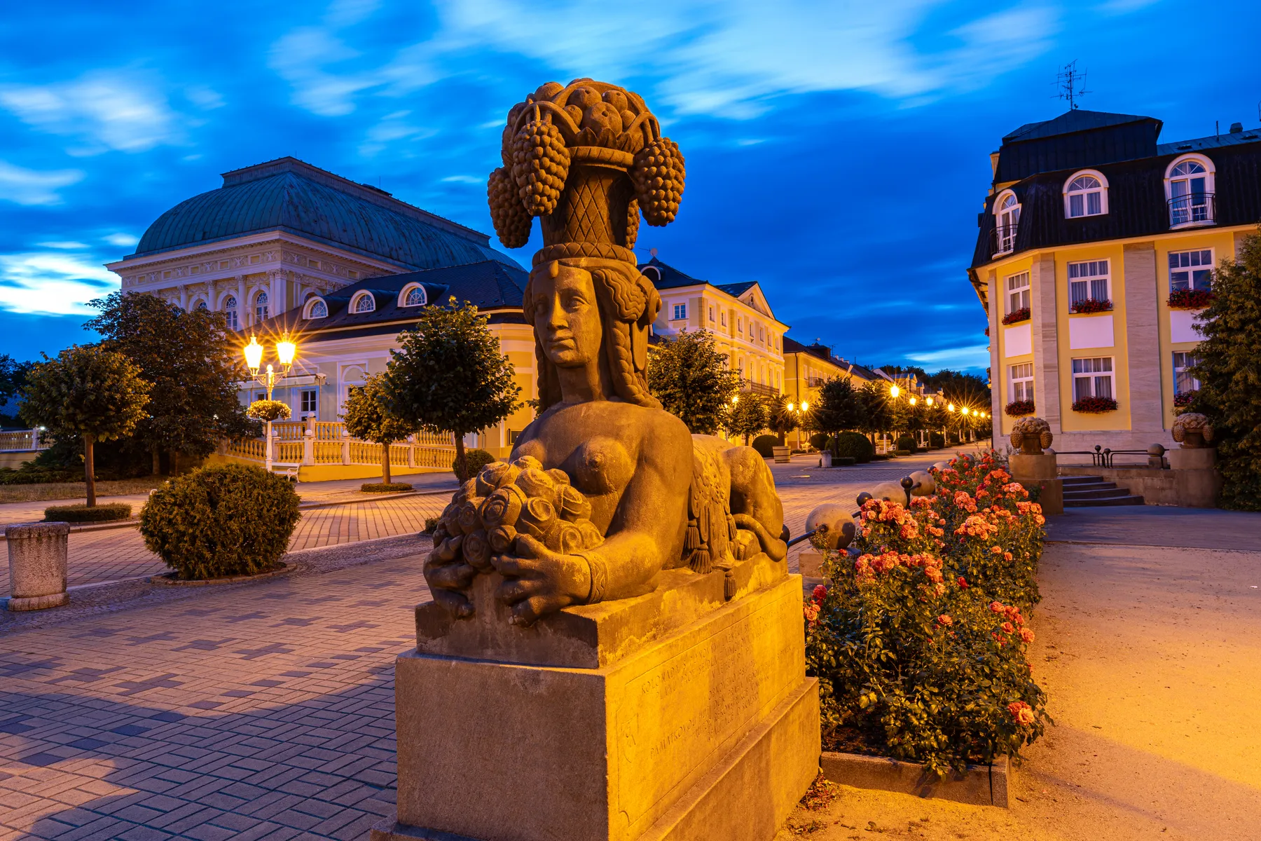 Eine steinerne Statue, die eine Frau mit einem Obstkorb auf dem Kopf darstellt, steht in der Abenddämmerung in einer ruhigen, blumengesäumten Straße von Franzensbad, mit verzierten Gebäuden und leuchtenden Straßenlampen im Hintergrund.