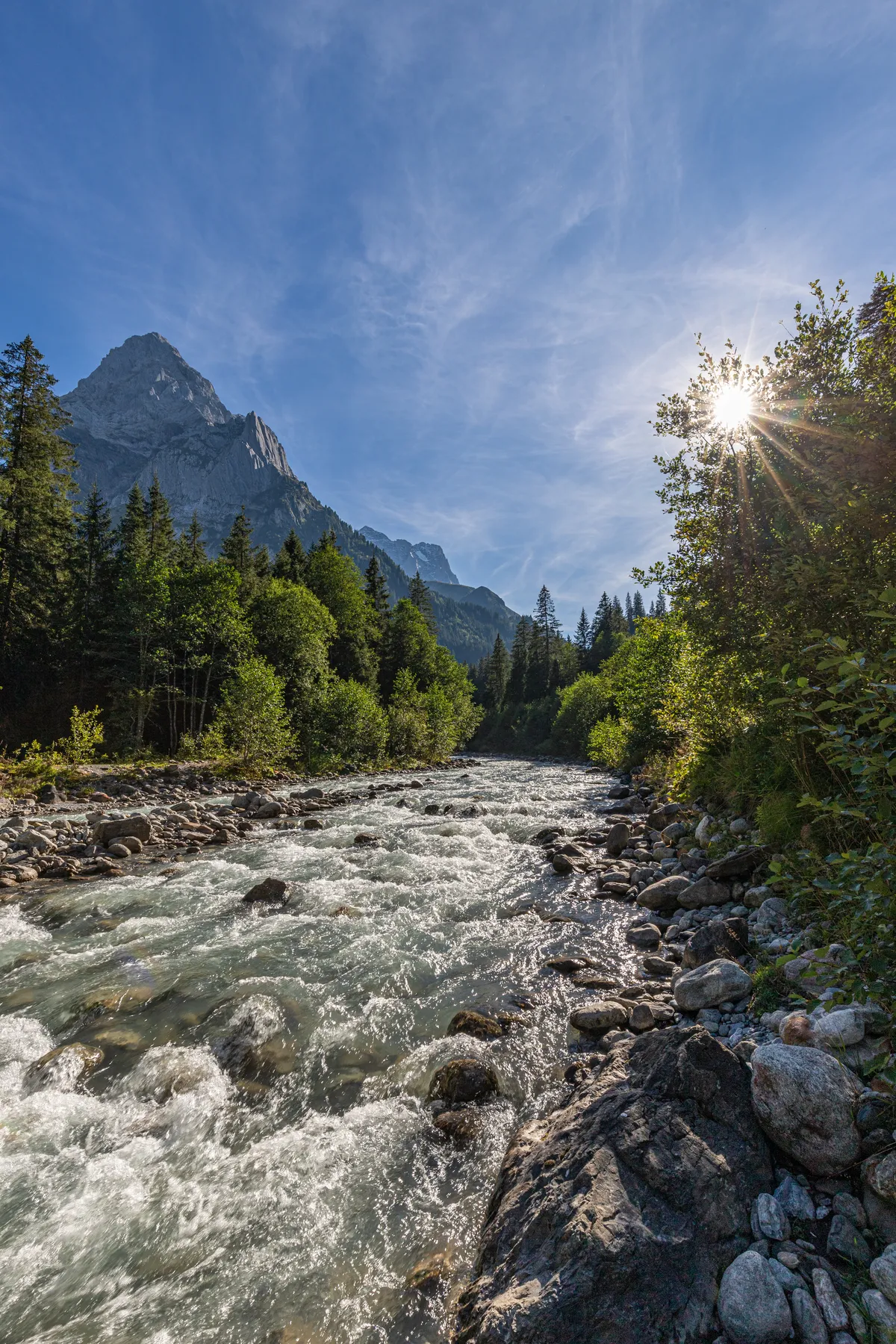 Ein klarer Fluss fließt über Felsen durch einen Wald mit hohen Bäumen. Im Hintergrund erhebt sich ein Berggipfel unter blauem Himmel, und das Sonnenlicht strömt durch die Bäume auf der rechten Seite und erzeugt einen Starburst-Effekt.