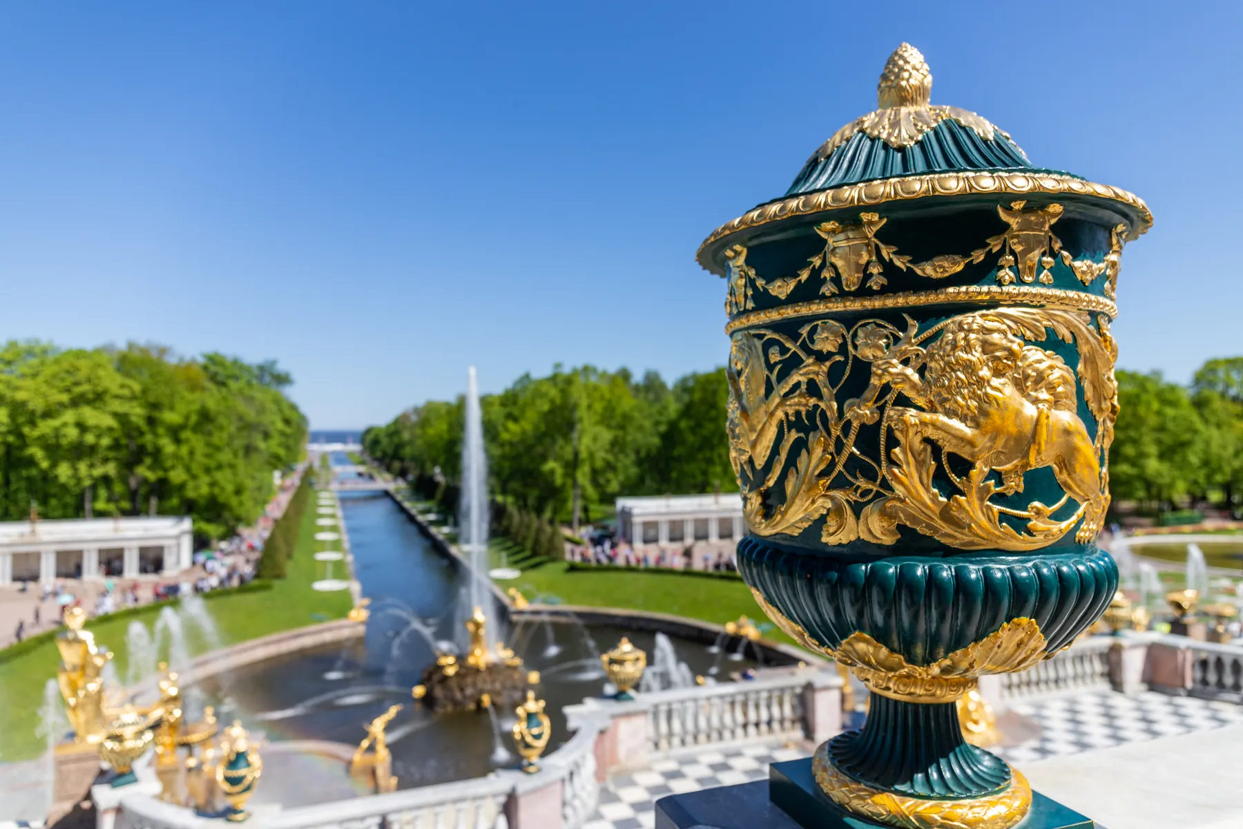 Im Vordergrund steht eine verzierte grün-goldene Urne mit einem Löwenmotiv, die sich an einem großen Kanal mit Brunnen, Statuen und gepflegten Gärten unter einem strahlend blauen Himmel befindet.