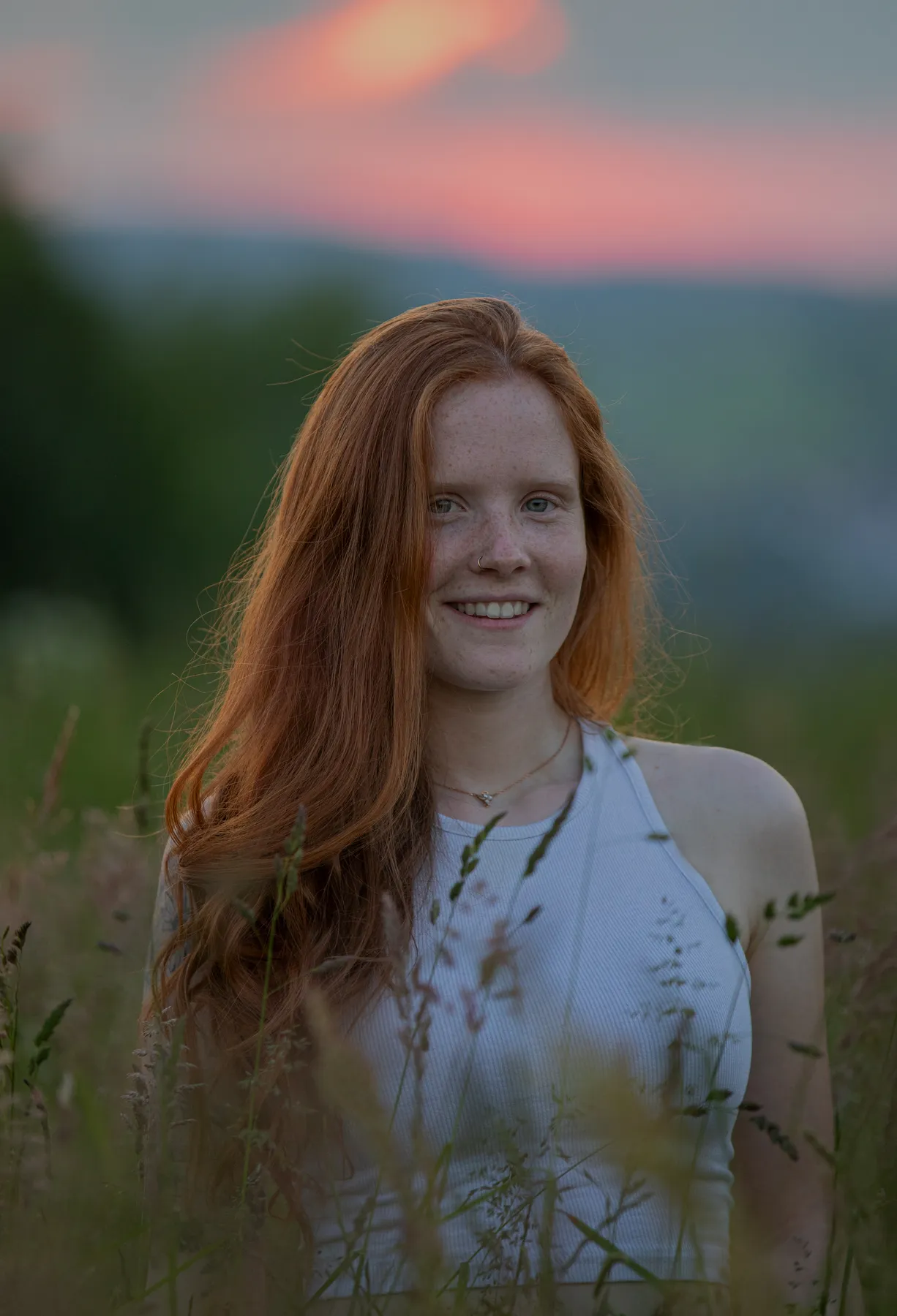 Eine junge Frau mit langen roten Haaren und Sommersprossen, die ein weißes ärmelloses Oberteil trägt, steht bei Sonnenuntergang auf einer Wiese mit einem verschwommenen rosa und blauen Himmel im Hintergrund. Sie lächelt sanft in die Kamera.