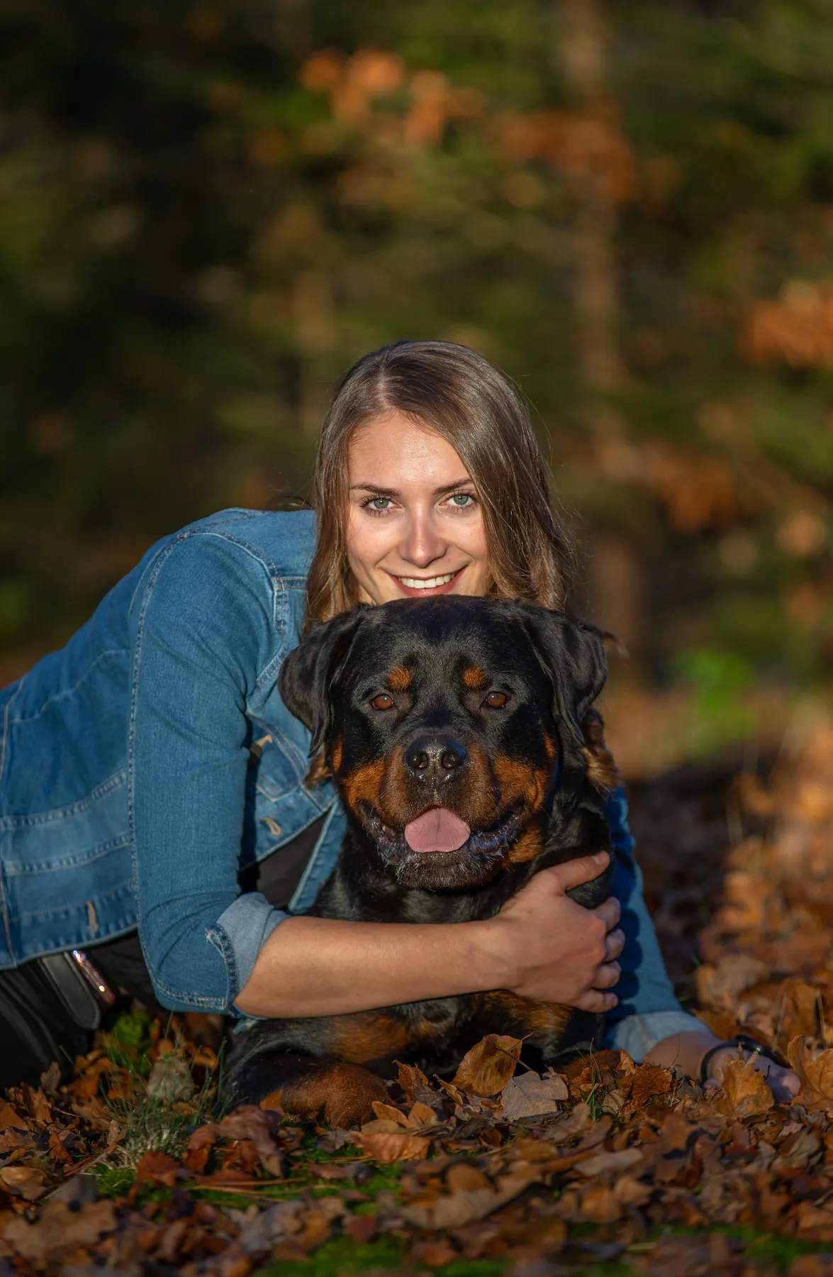 Eine lächelnde Frau in Jeansjacke umarmt einen Rottweiler, während sie auf Herbstblättern in einem sonnenbeschienenen Wald liegt. Sie schaut über seinem Kopf hinweg, der Rottweiler schaut gelassen und hat leicht die Zunge herausgestreckt.