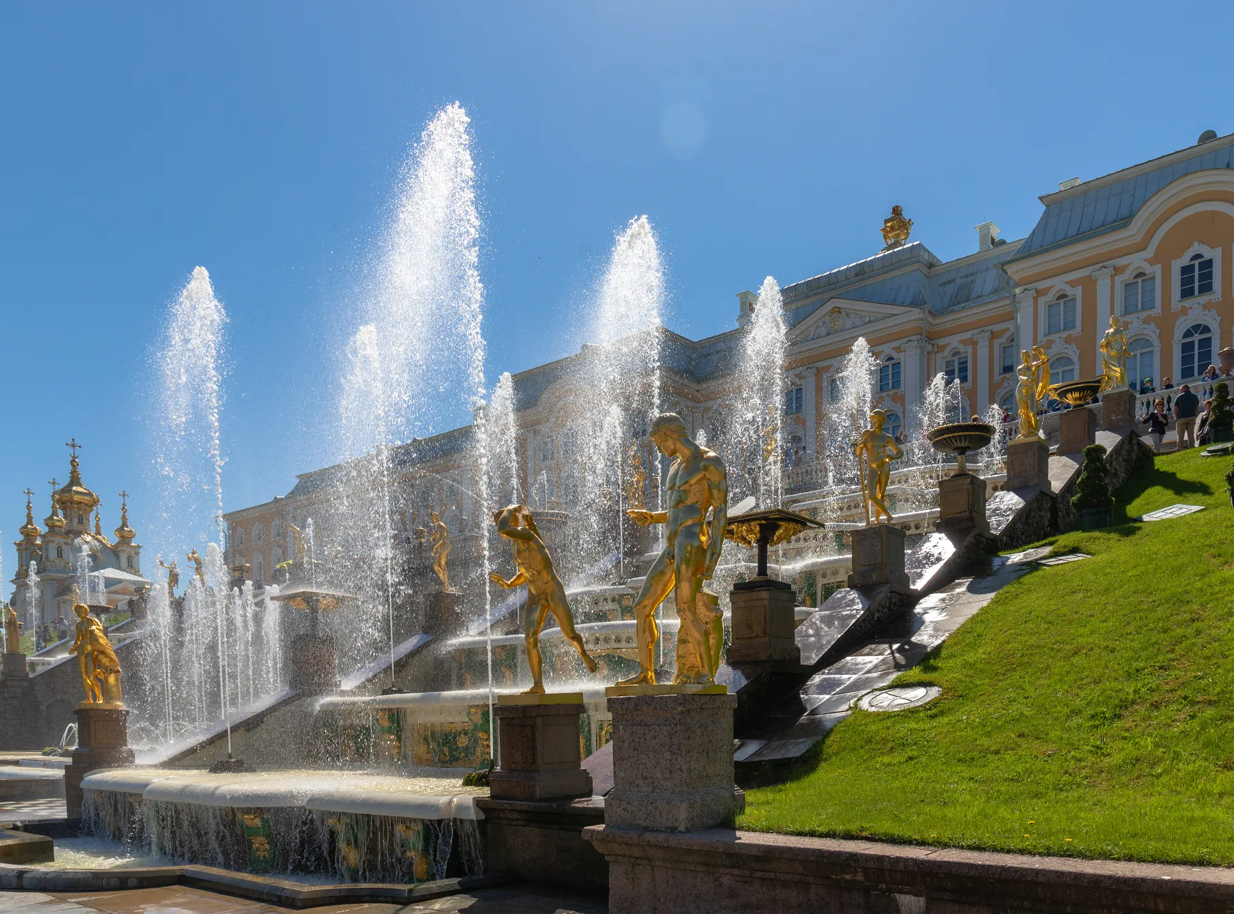 Große goldene Statuen und Springbrunnen vor dem prächtigen Peterhof-Palast in St.-Petersburg, unter blauem Himmel, mit gepflegten grünen Rasenflächen auf beiden Seiten. Im Hintergrund historische Gebäude
