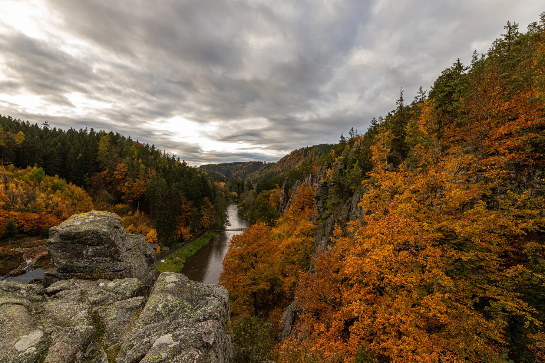 Die Eger schlängelt sich durch ein bewaldetes Tal in Tschechien mit Herbstlaub in leuchtenden Orange- und Gelbtönen, flankiert von felsigen Klippen und immergrünen Bäumen unter einem dramatischen, bewölkten Himmel.