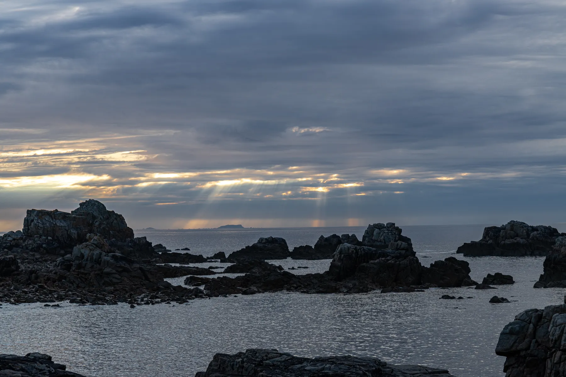 Landschaftsfotografie eines Zerklüfteten Küstenabschnitt der bretonischen Küste bei Ebbe. Im Vordergrund ragen die Felsformationen aus dem Wasser das ruhig und leicht wellig ist. Derr Himmel ist bedeckt aber durch die Wolkenlücken brechen die Sonnenstrahlen