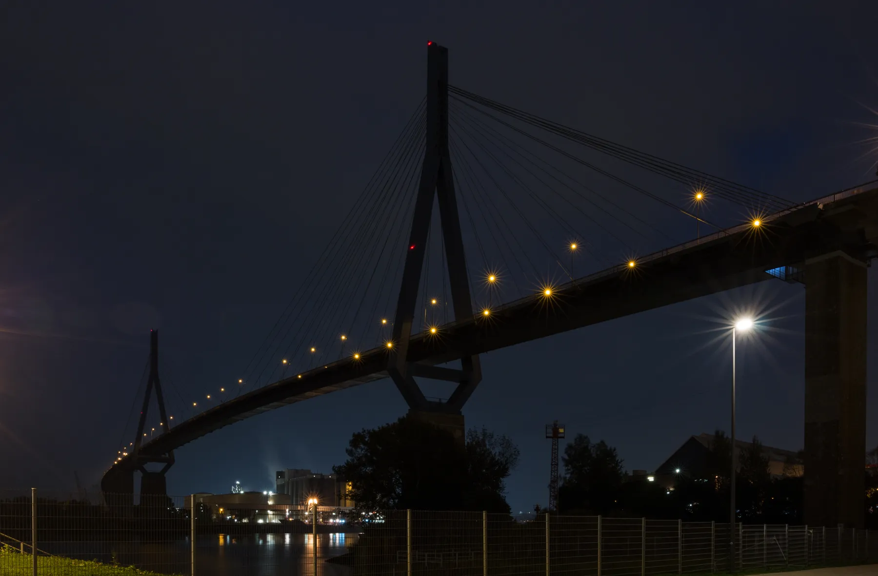 Landschaftsfotografie der mächtigen Köhlbrandbrücke bei Nacht mit beleuchteten Straßenlaternen. Im Hintergrund ist eine Siloanlage zu sehen deren Lichter im wasser sich spiegeln