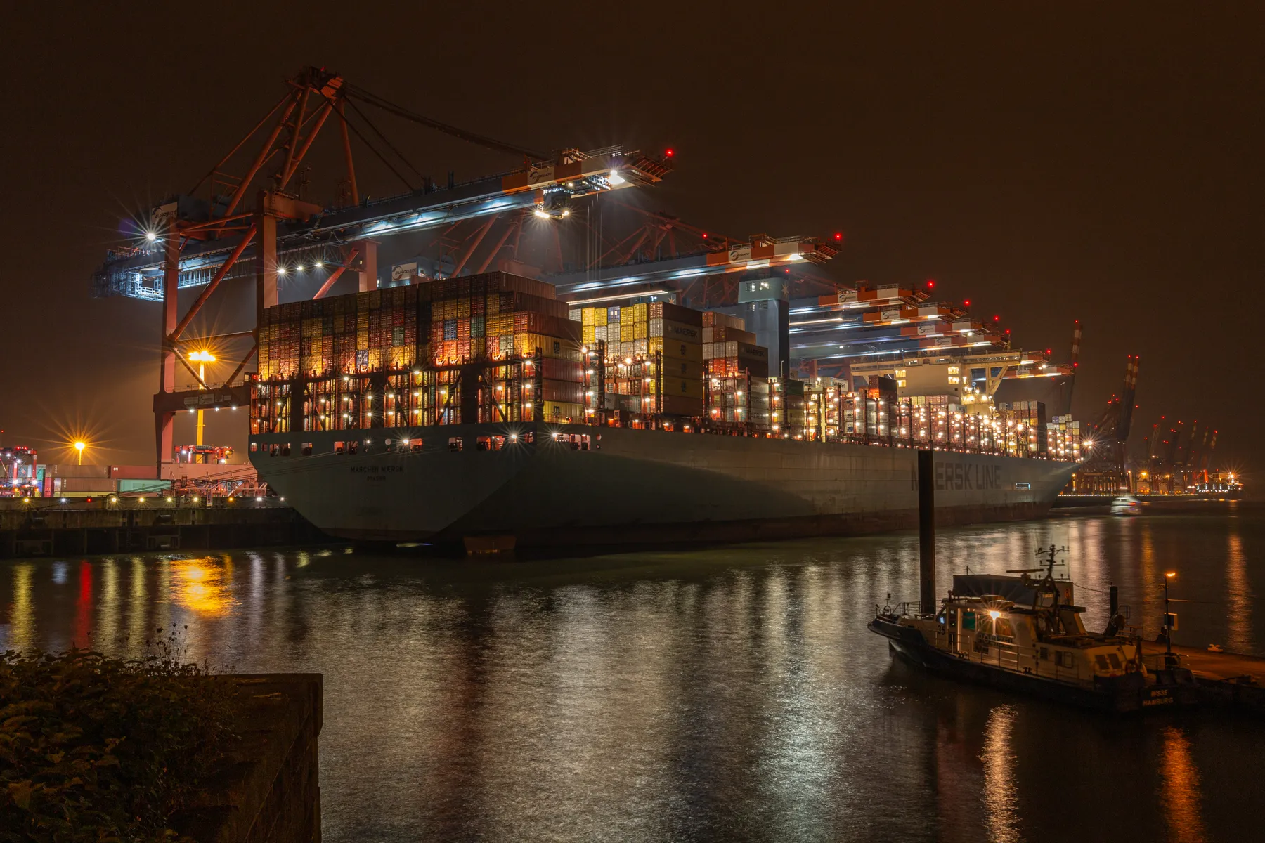 Ein riesiges Containerschiff liegt am Burchardkai und wird gerade beladen. Die Szenerie ist bei nacht und alles wird erhellt von vielen Lichtern des Schiffes, der Kaianlage und der Portalkräne welche über dem Schiff zu sehen sind