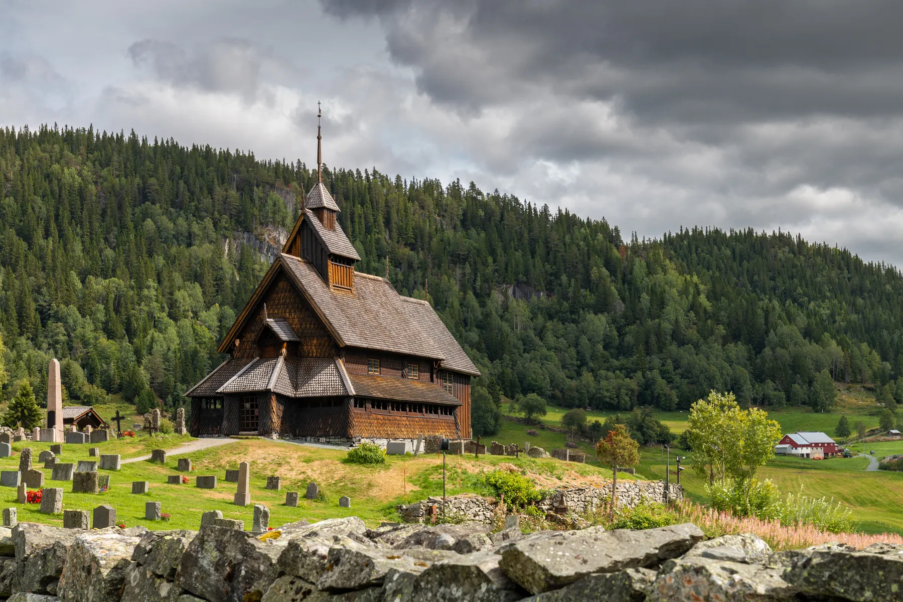 Landschaftsfotografie der Stabkirche Eidsvoll zwischen Grabsteinen, umgeben von grünen Hügeln und dichtem Wald unter einem bewölkten Himmel, mit einer Steinmauer im Vordergrund und ein paar entfernten Gebäuden im Hintergrund.