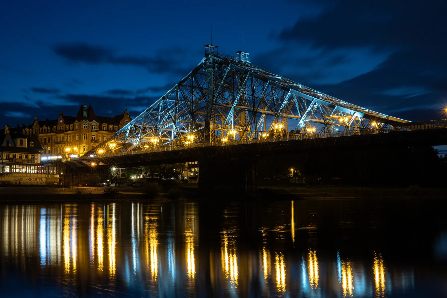 Eine große, beleuchtete Stahlbrücke, das Blaue Wunder in Dresden, überspannt die ruhige nächtliche Elbe, dessen goldene Lichter sich im Wasser spiegeln. Auf der linken Seite sind Gebäude mit leuchtenden Fenstern unter einem tiefblauen Himmel zu sehen.