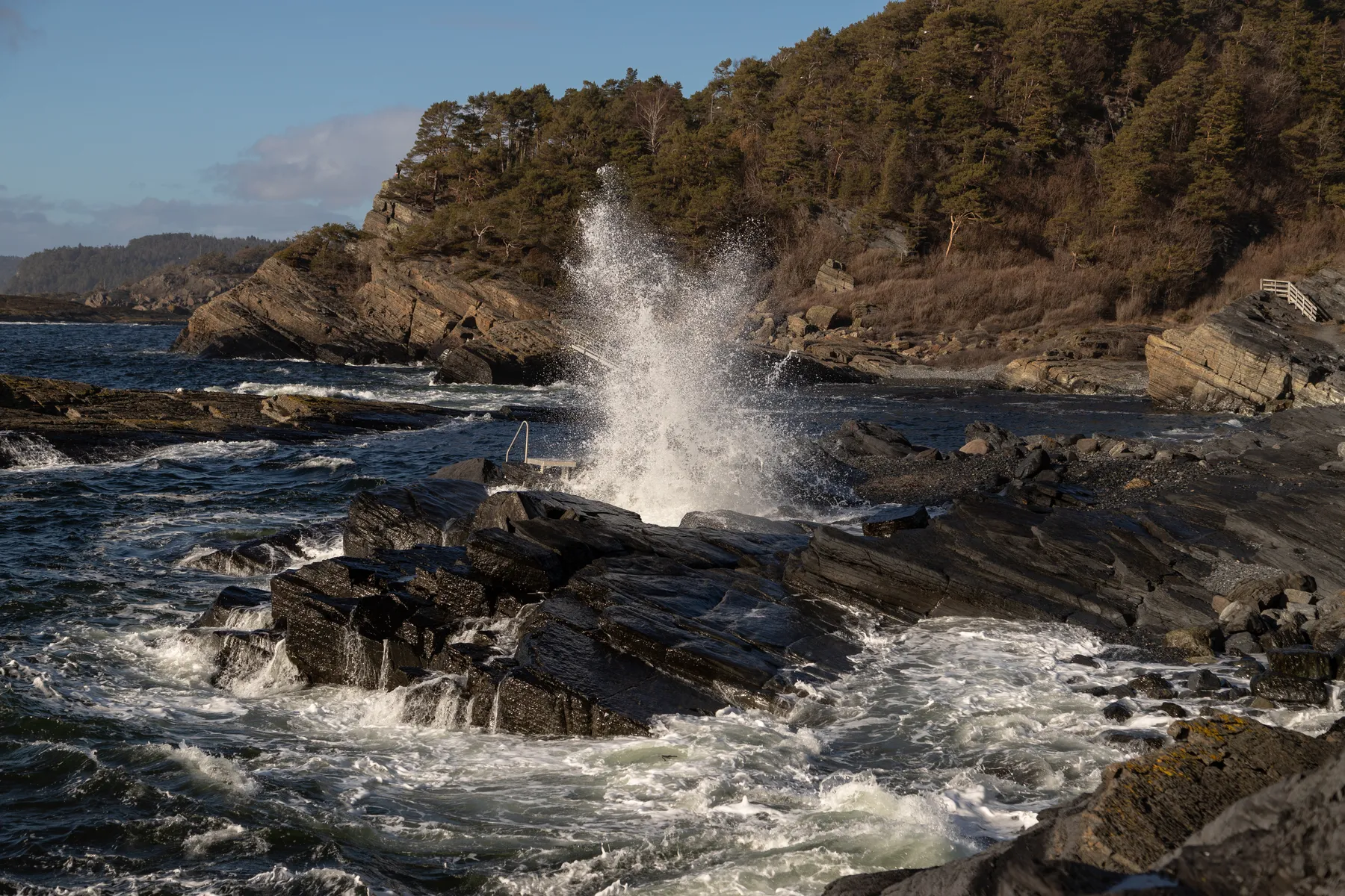 An der zerklüfteten Küste von Langesund schlagen die Wellen dramatisch gegen dunkle Felsen und lassen eine Gischt aufsteigen. Im Hintergrund erhebt sich ein bewaldeter Hügel unter einem teilweise bewölkten blauen Himmel.