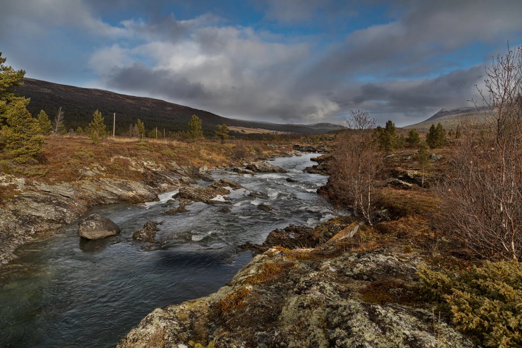 Ein felsiger Fluss schlängelt sich durch eine zerklüftete Landschaft im norwegischen Gebirge mit spärlichen Bäumen und Sträuchern unter einem wolkenverhangenen Himmel, mit fernen Hügeln und Bergen im Hintergrund.