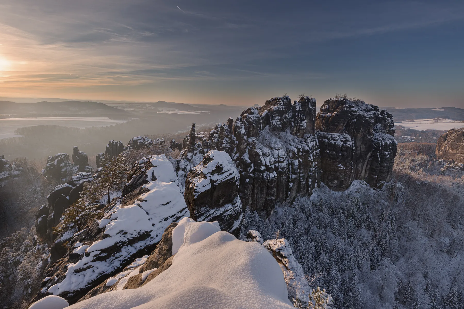 Die schneebedeckten Felsformationen der Schrammsteine erheben sich bei Sonnenuntergang über einem frostigen Wald im Elbsandsteingebirge, mit einem sanften Leuchten am Horizont und eisigen Bäumen darunter.