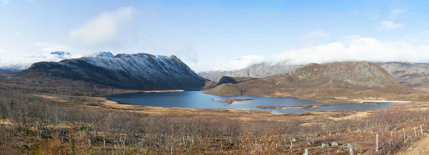 Panoramablick auf Berge in der Valdresflye in Norwegen, einen ruhigen See inmitten schneebedeckter Berge unter einem teilweise bewölkten blauen Himmel, mit spärlichem Herbstlaub und kahlen Bäumen im Vordergrund.