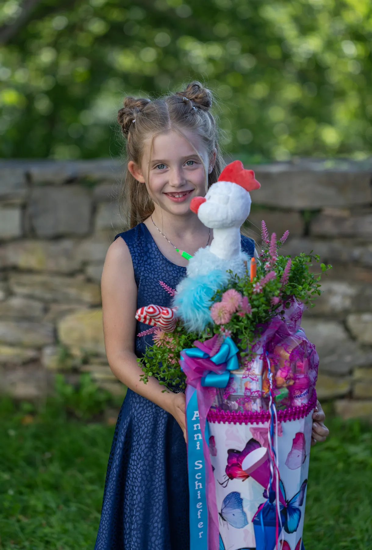 Ein junges Mädchen in einem blauen Kleid hält lächelnd die Schulanfang-Zuckertüte - eine große, bunte, mit Blumen und einem Plüschhuhn verzierte Tüte - in der Hand, während sie im Freien an einer Steinmauer und im Grünen steht.