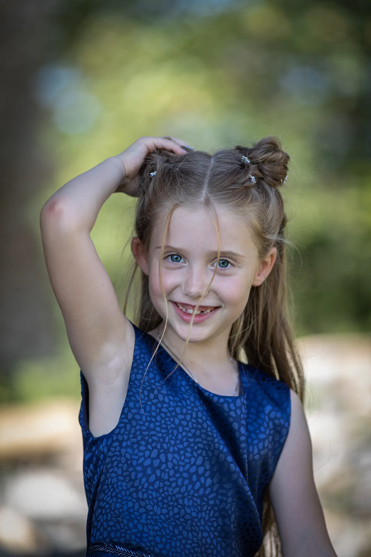 Ein junges Mädchen mit langen blonden Haaren, die zu zwei Dutts frisiert sind, sie trägt ein blau gemustertes Kleid und steht im Freien, lächelt und hält eine Hand auf ihren Kopf. Der Hintergrund ist unscharf mit Grünzeug.