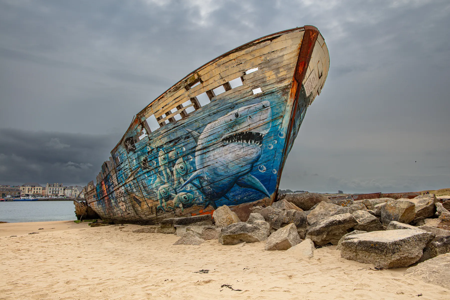 Ein altes, verwittertes Schiffswrack liegt am Sandstrand in der Nähe des Flusses Étel in der Südbretagne. Seine Seite ist mit einem lebhaften Wandgemälde eines großen Hais und Fisches verziert, vor dem Wrack liegen Felsen und der Himmel ist wolkenverhangen. Im Hintergrund am jenseitigen Ufer sind Häuser zu sehen.