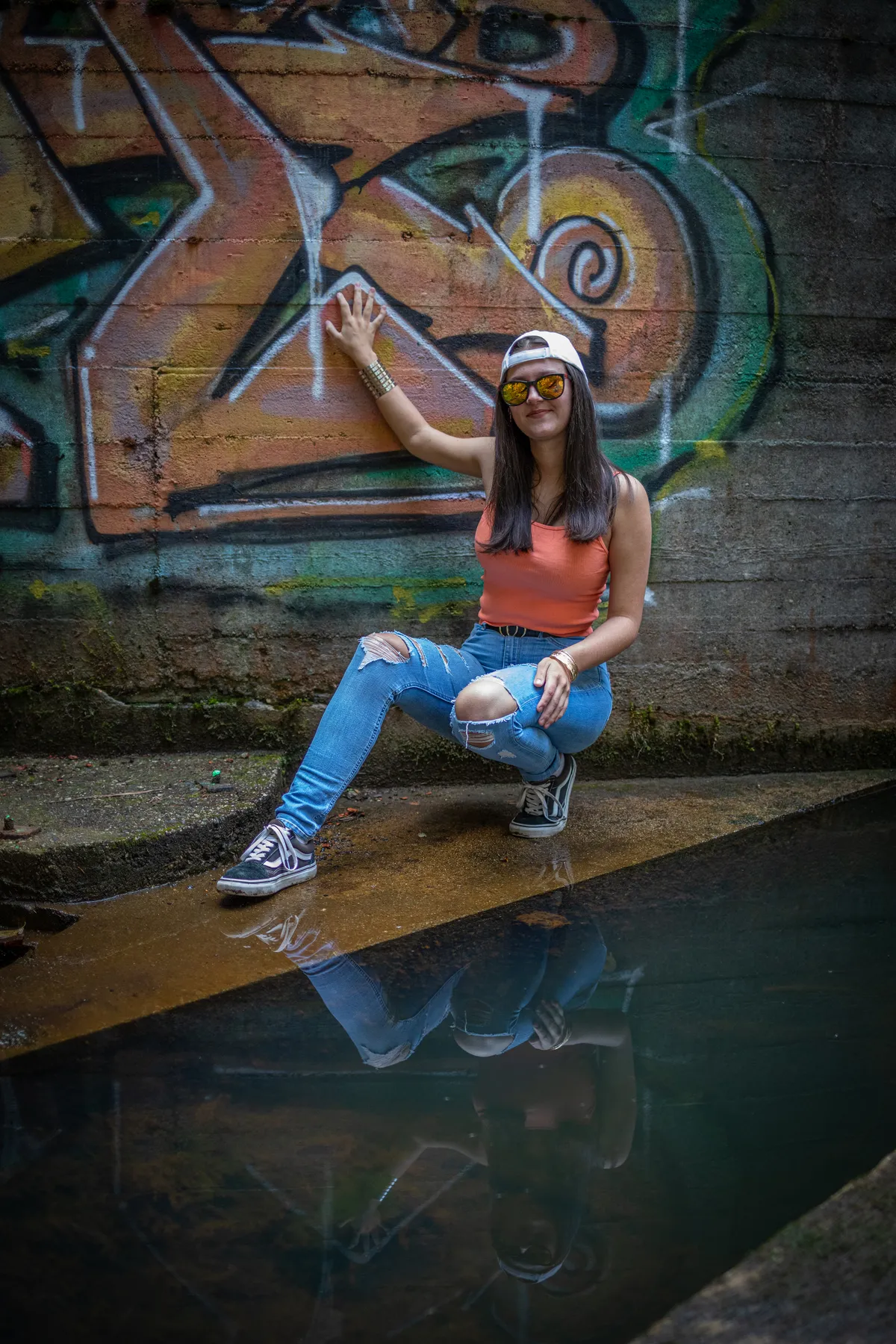 Eine Frau in einem korallenroten Tank-Top, zerrissenen Jeans und Sonnenbrille posiert an einer mit Graffiti beschmierten Wand und kniet neben einem flachen Wasserbecken, in dem sich ihr Bild spiegelt.