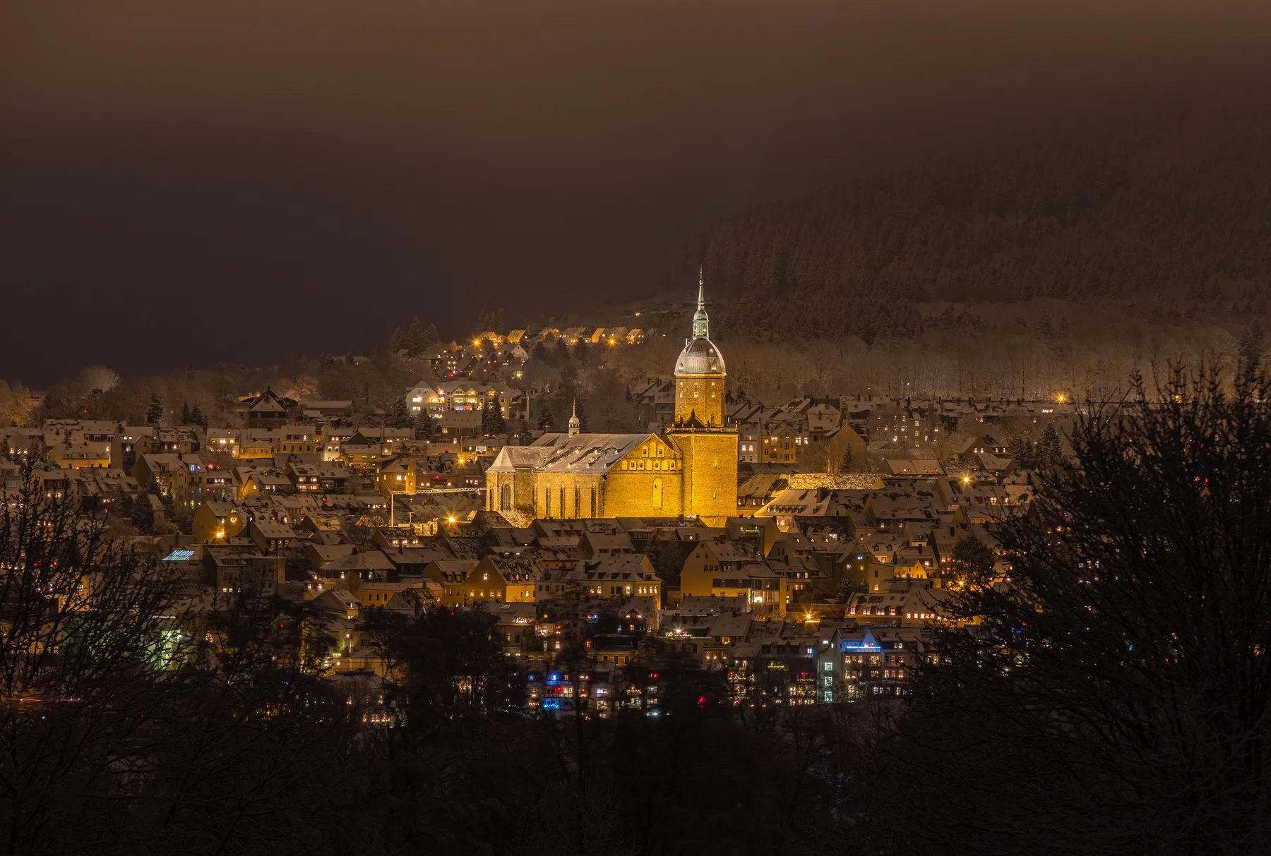 Annenkirche in Annaberg Buchholz im winterlichen Abendlicht, untermalt durch die vielen Fensterbeleuchtungen. Die Dächer und sind schneebedeckt, der Wald im Hintergrund verschwimmt mit der Dunkelheit.