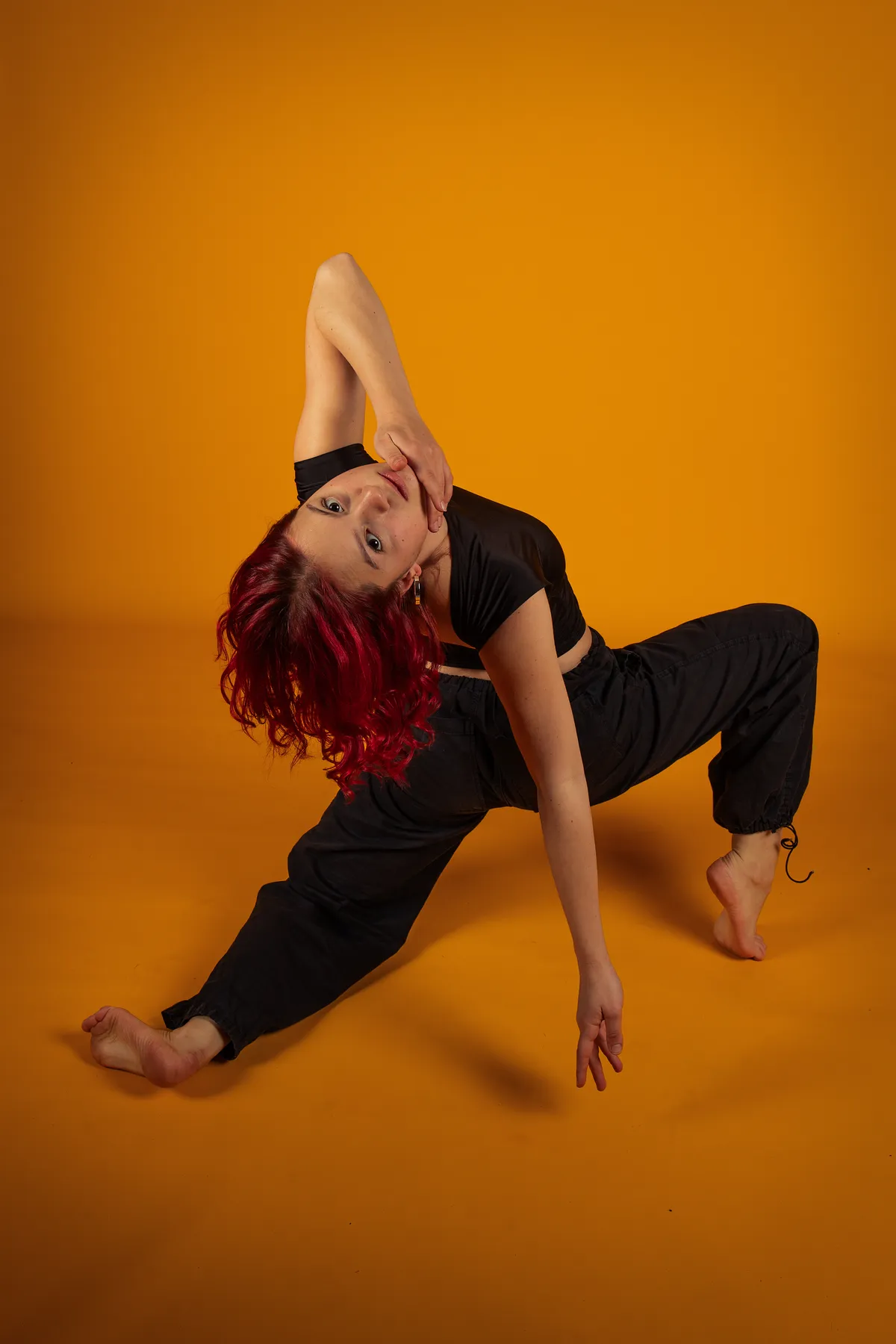 Eine junge Frau mit roten Haaren, bekleidet mit einer schwarzen Hose und einem schwarzen Crop-Top, posiert barfuß in einer dynamischen, nach hinten gebeugten Pose vor einem orangefarbenen Hintergrund.