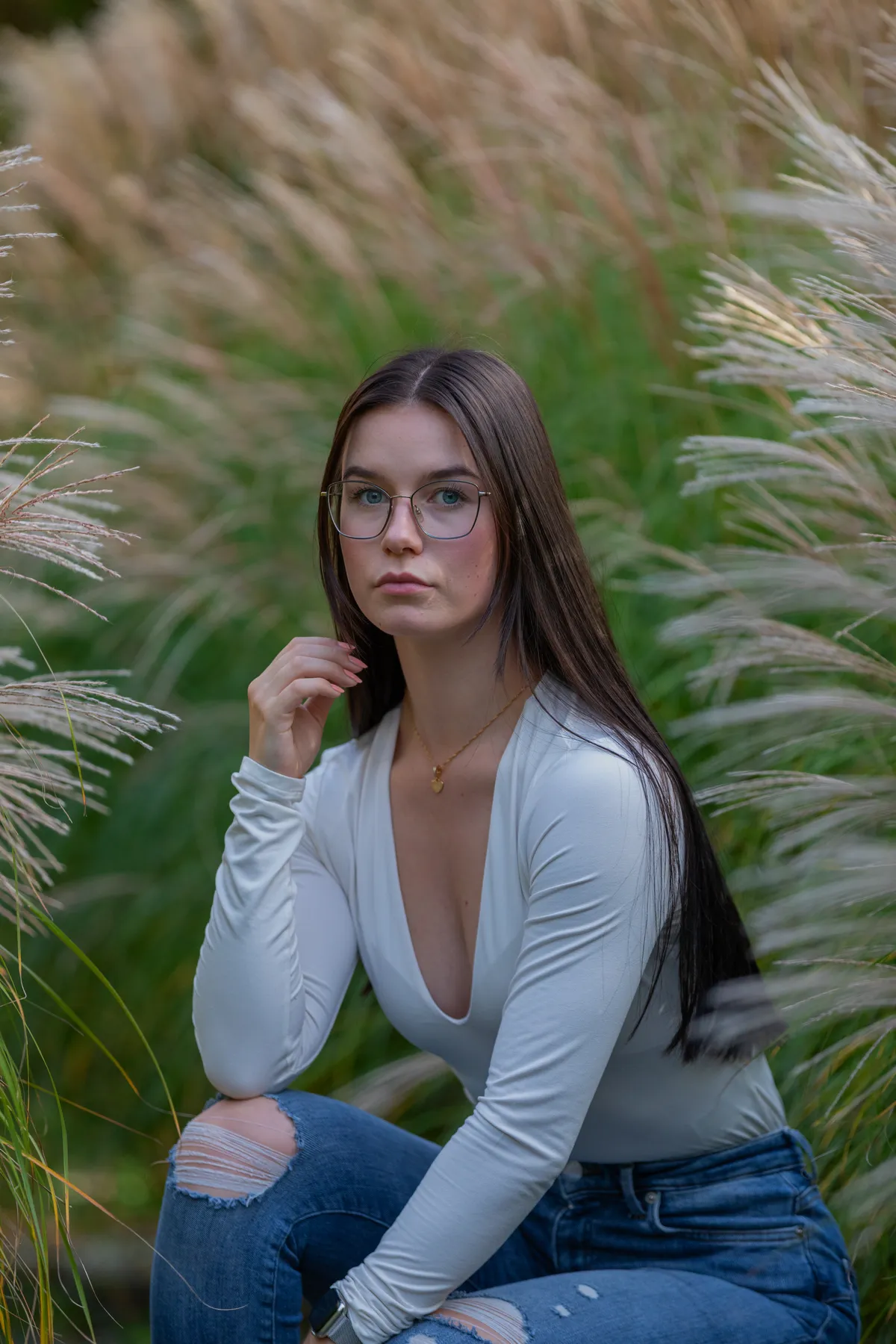 Eine junge Frau mit langem, glattem braunem Haar und Brille sitzt im hohen Gras, trägt ein weißes, langärmeliges Oberteil mit tiefem Ausschnitt und zerrissene Bluejeans und schaut mit neutralem Blick direkt in die Kamera.
