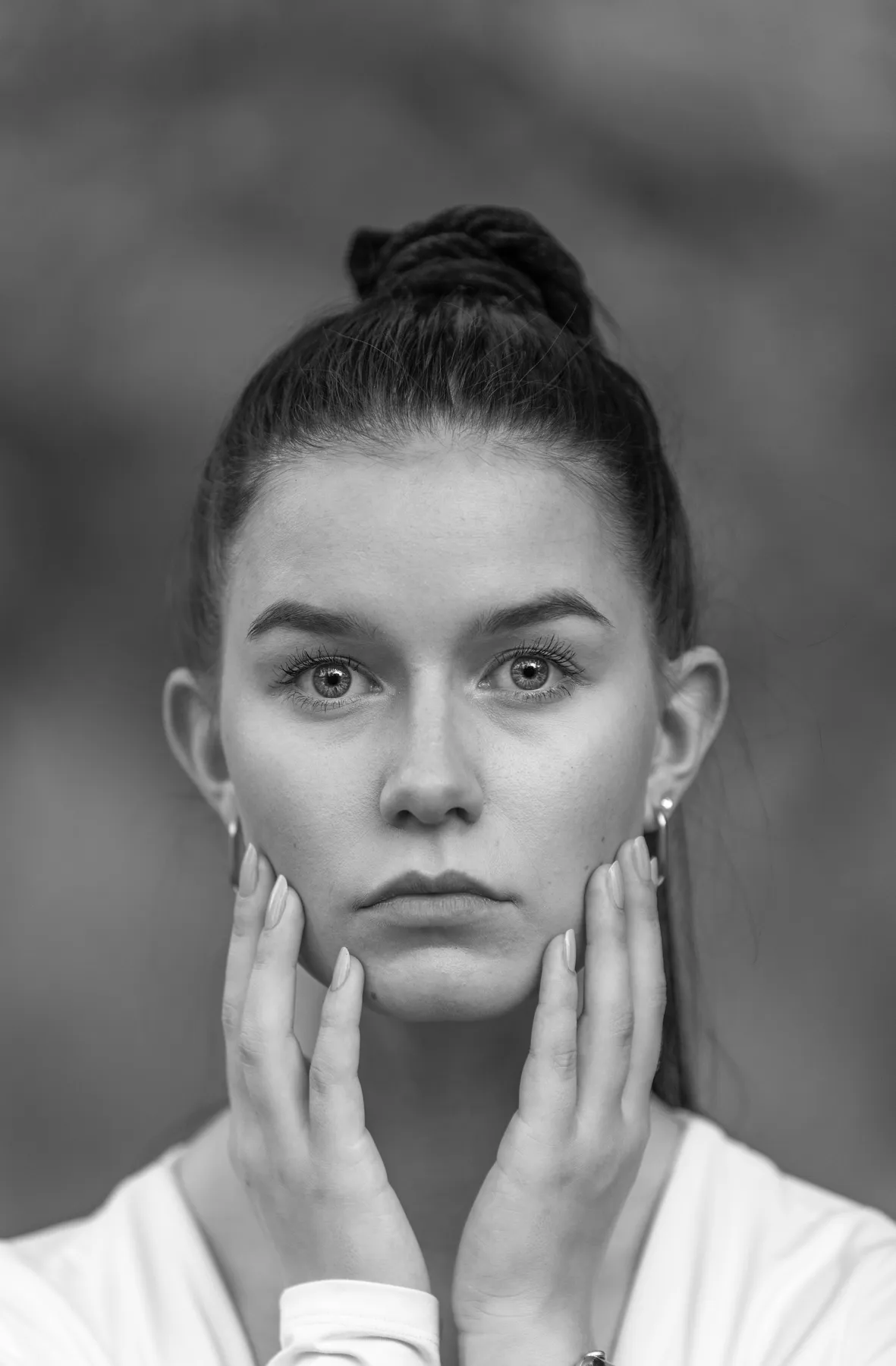 Eine junge Frau mit dunklem Haar in einem hochgesteckten Dutt blickt mit neutralem Ausdruck direkt in die Kamera, beide Hände berühren sanft ihre Wangen. Der Hintergrund ist unscharf.
