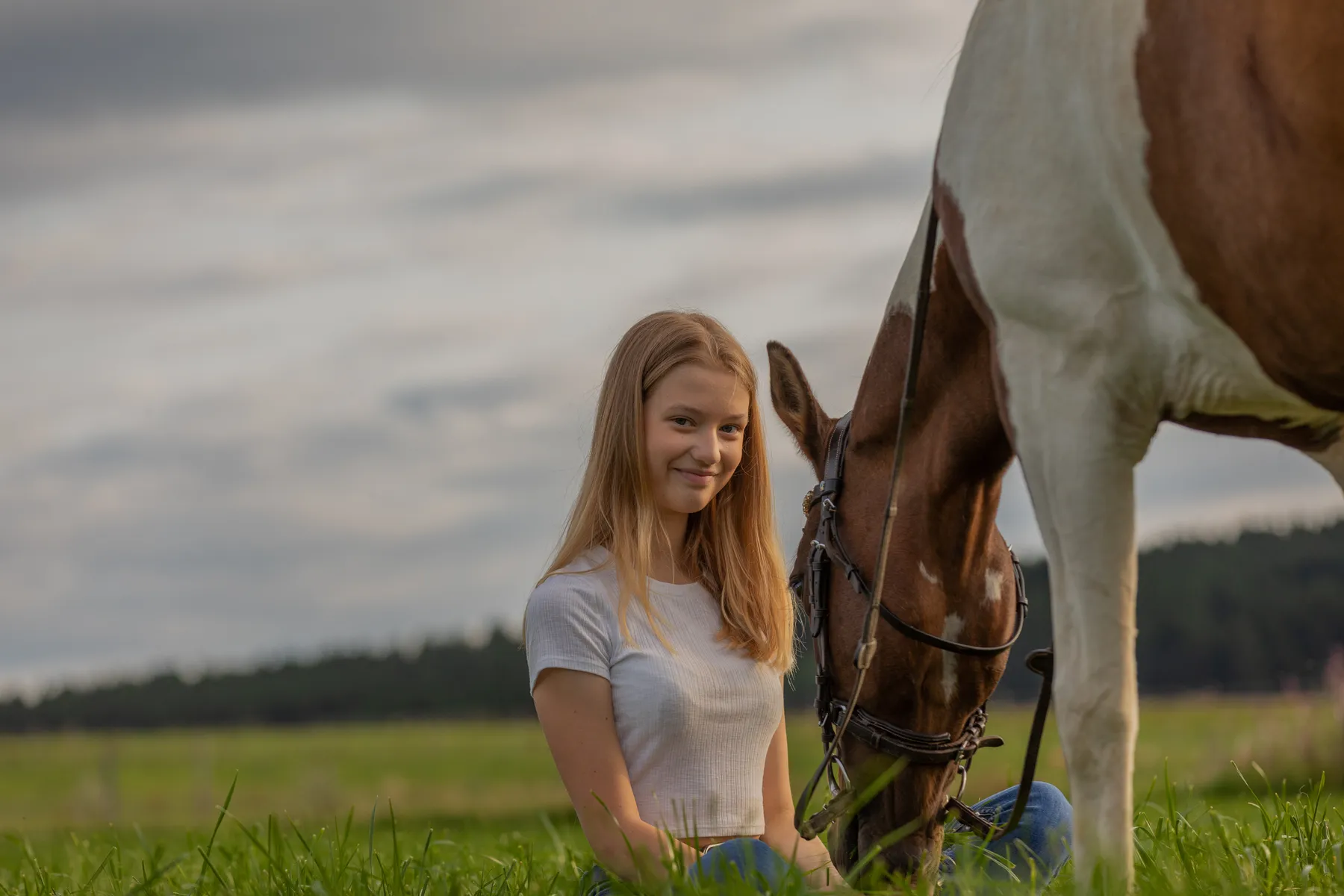 Eine junge Frau mit langen blonden Haaren, die ein weißes T-Shirt trägt, sitzt im Gras neben einem braun-weißen Pferd, das weidet. Im Hintergrund ist ein Feld mit Bäumen unter einem bewölkten Himmel zu sehen.
