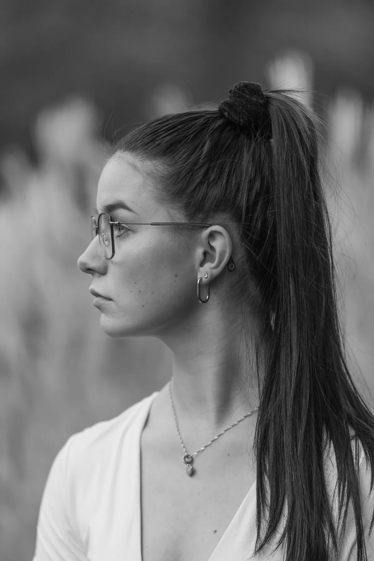 Schwarz-Weiß-Foto einer jungen Frau mit langen, dunklen Haaren in einem hohen Pferdeschwanz, die eine Brille, Ohrringe und eine Halskette trägt, dargestellt im Profil vor einem unscharfen Hintergrund im Freien.
