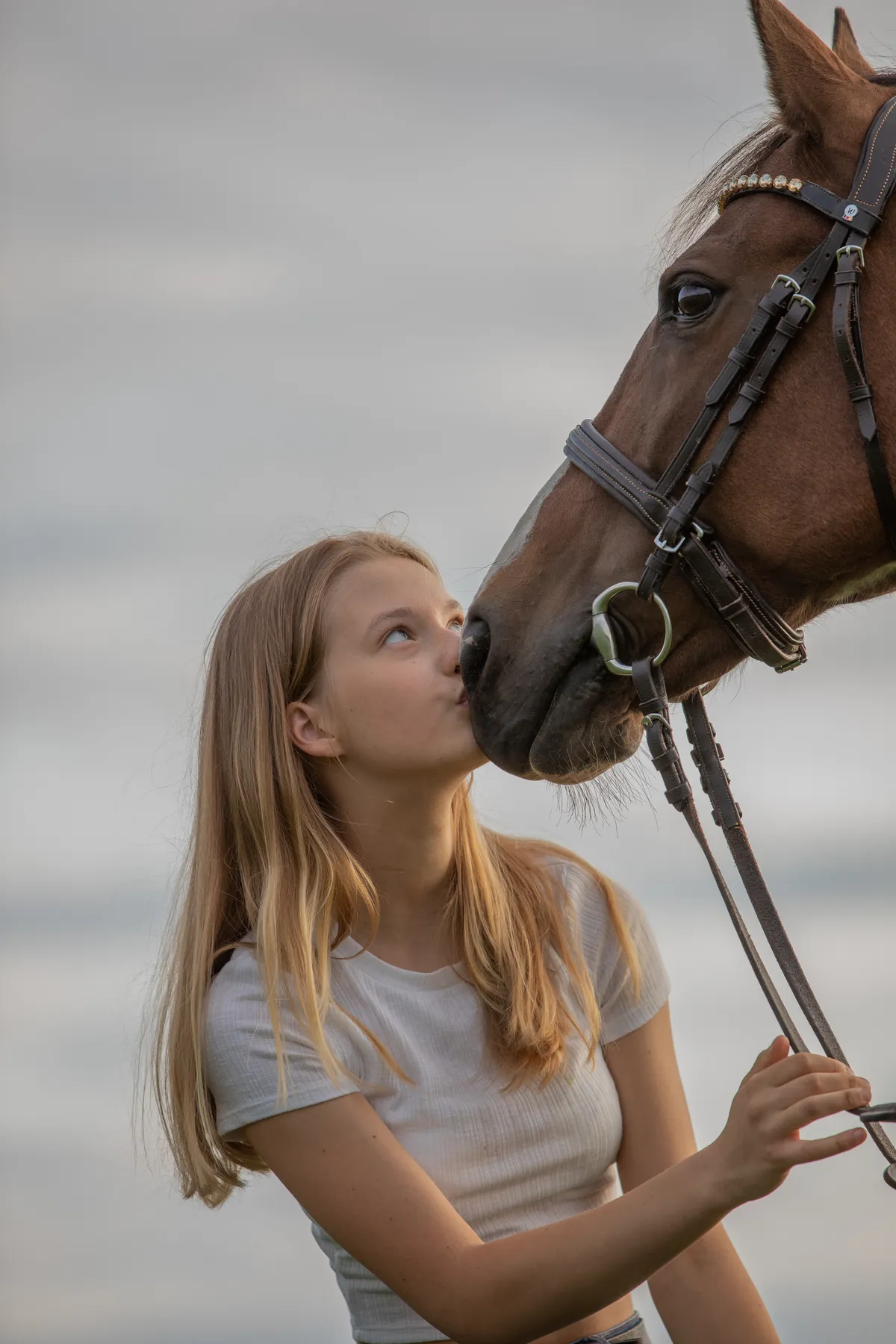 Eine junge Frau mit langen blonden Haaren und einem kurzärmeligen hellen Hemd steht im Freien, hält sanft die Zügel eines Pferdes und küsst die Nase des Pferdes. Der Hintergrund ist unscharf und neutral.