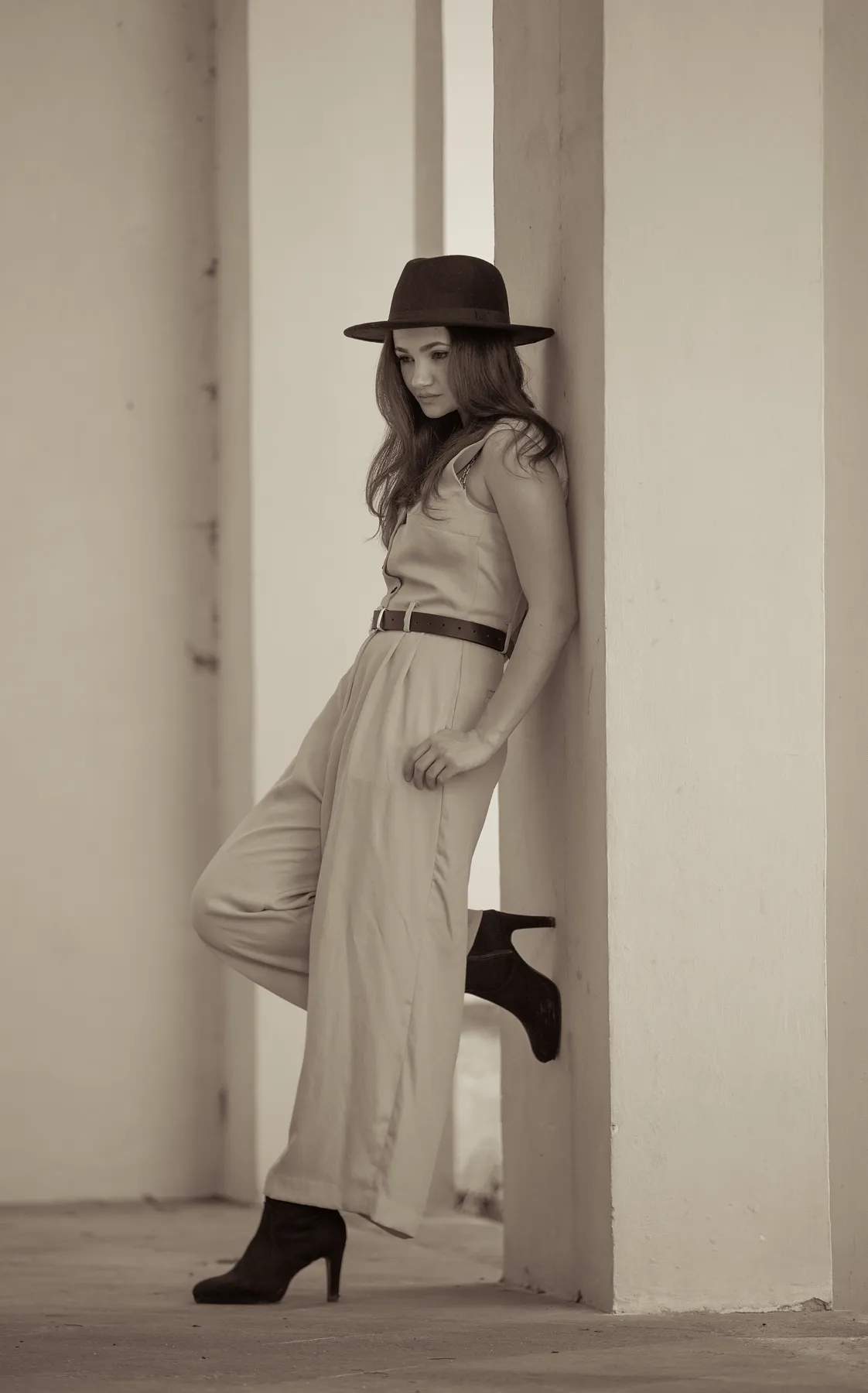 Eine Frau mit einem breitkrempigen Hut, einem ärmellosen Overall und hochhackigen Stiefeln lehnt an einer hohen, hellen Wand und blickt mit einem angewinkelten Knie nach unten. Das Bild ist in Sepiatönen gehalten. Das Bild vermittelt Eleganz