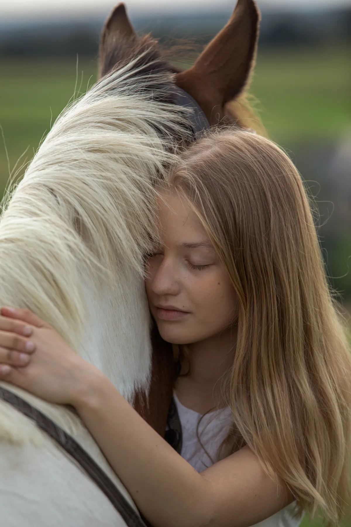Ein junges Mädchen mit langen blonden Haaren umarmt ein weiße/braunes Pferd, wobei sie ihre Wange mit geschlossenen Augen an den Hals des Pferdes lehnt, in einer Umgebung im Freien mit unscharfem grünen Gras im Hintergrund.