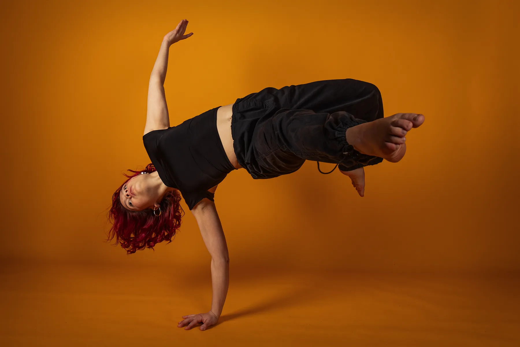 Eine Person mit roten Haaren, die ein schwarzes Crop-Top und eine Hose trägt, balanciert auf einer Hand in einer Breakdance-Pose vor einem orangefarbenen Hintergrund.