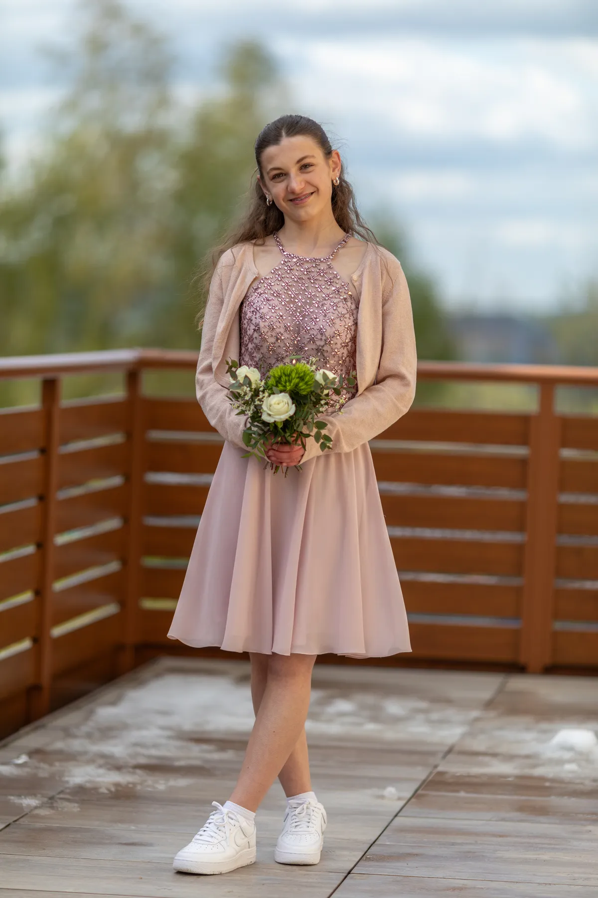 Ein junges Mädchen lächelt auf einer hölzernen Plattform. Es trägt ein hellrosa Kleid zu seiner Konfirmation, eine passende Strickjacke und weiße Turnschuhe und hält einen kleinen Blumenstrauß in der Hand. Im Hintergrund sind Bäume und ein bewölkter Himmel zu sehen.