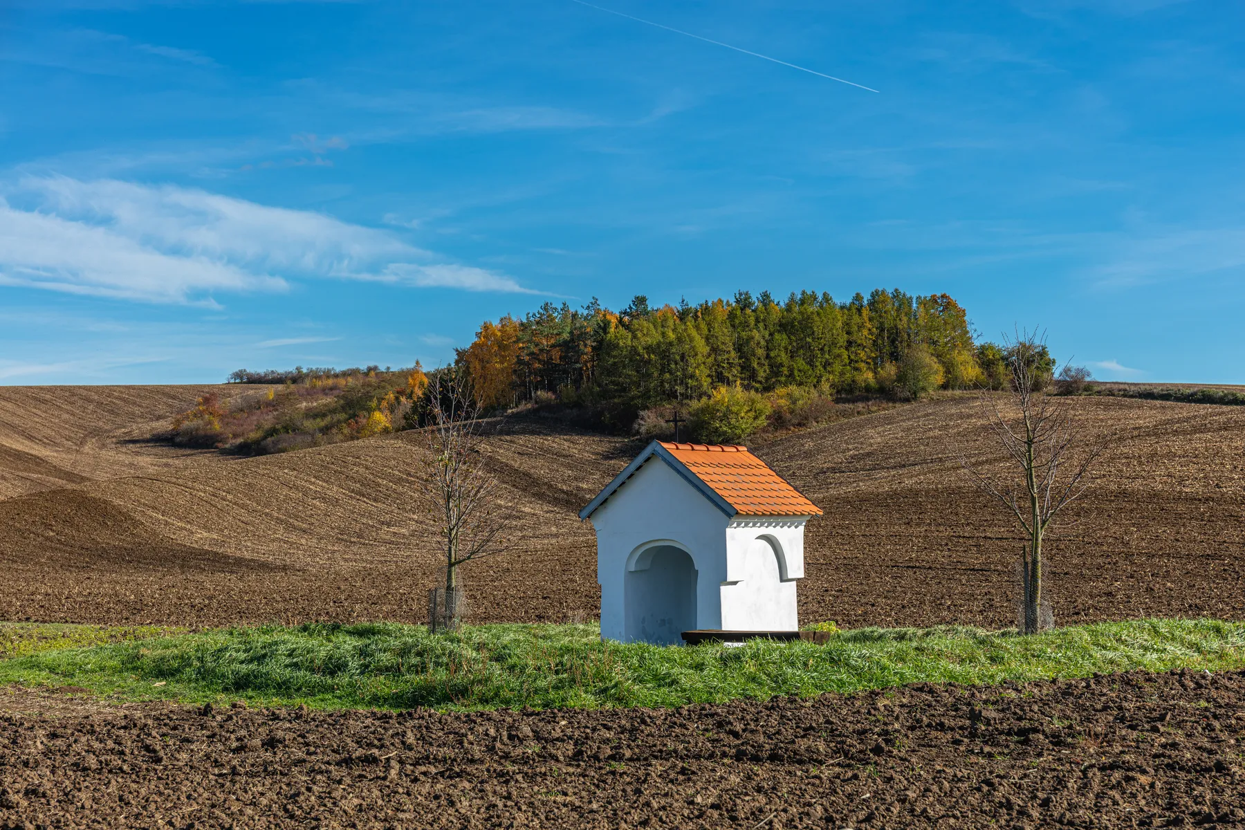 Eine kleine weiße Kapelle mit rotem Dach steht auf grünem Gras zwischen Bäumen, eingerahmt von gepflügten Feldern und Hügeln unter einem blauen Himmel in der Mährischen Toskana.