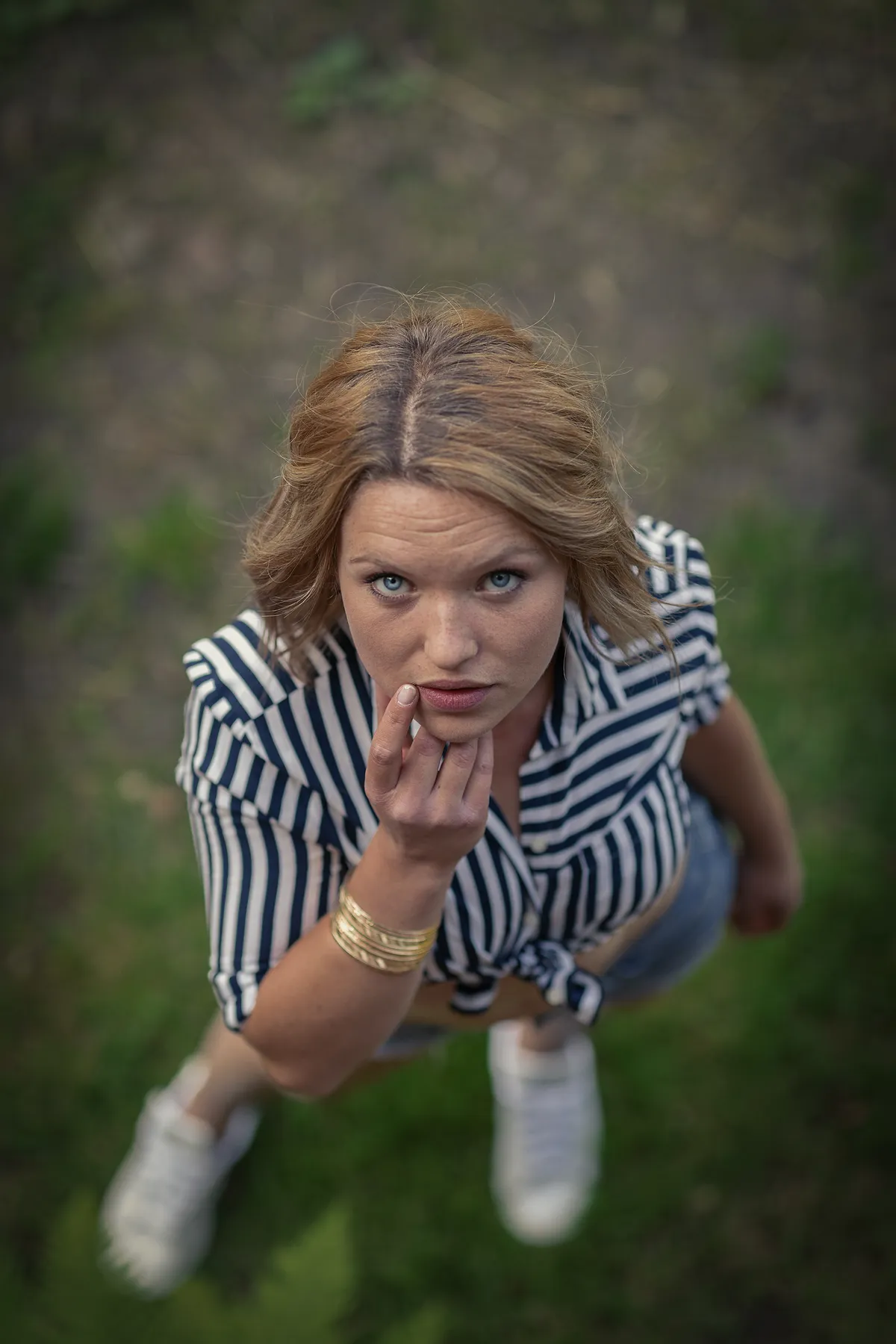 Eine Frau mit blonden Haaren, einem schwarz-weiß gestreiften Hemd, Jeansshorts und weißen Schuhen steht im Gras und blickt in die Kamera, während sie mit der Hand ihr Kinn berührt. Das Foto ist von oben aufgenommen.