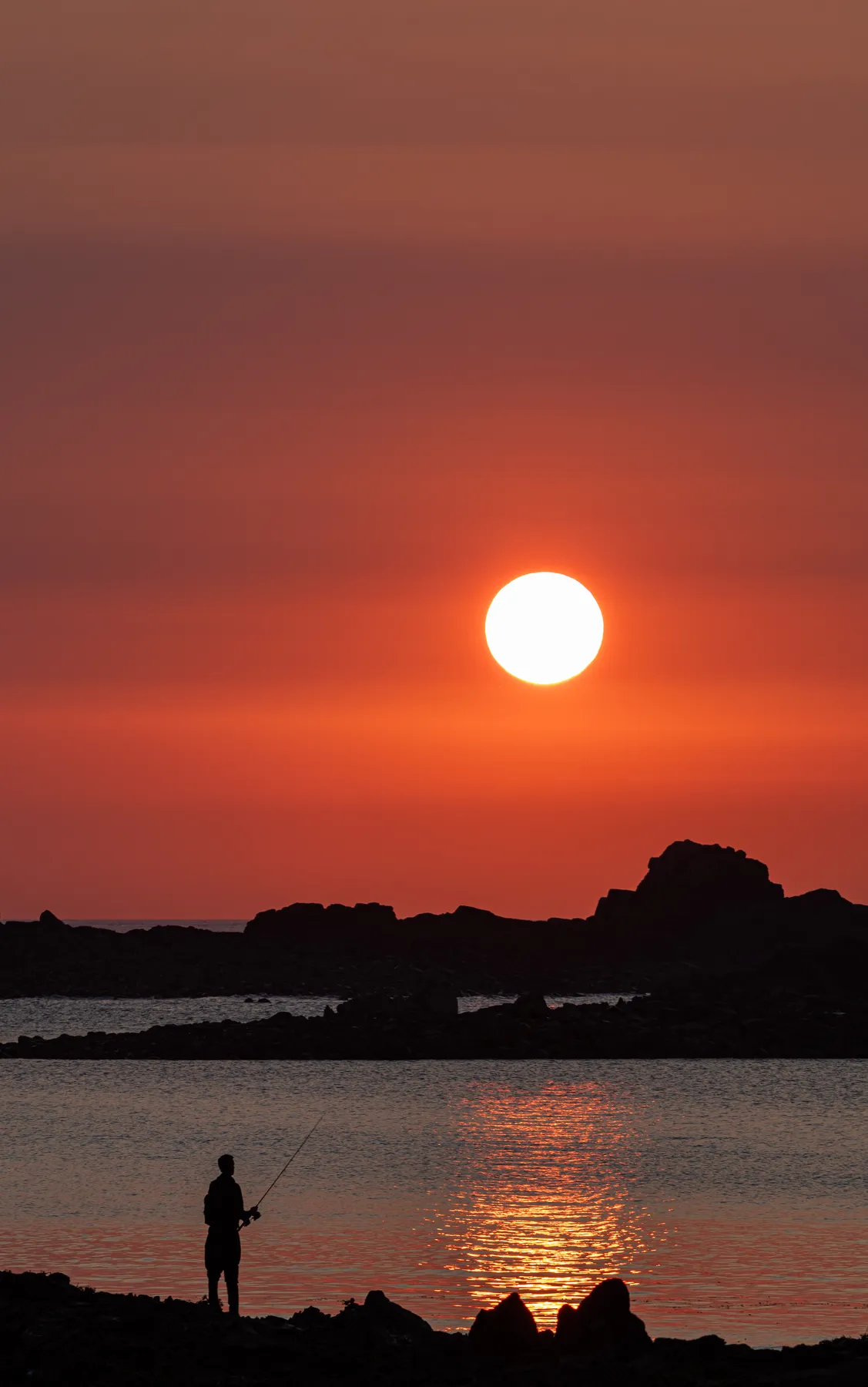 Landschaftsfotografie eines atemberaubenden Sonnenuntergangs über dem Meer. Im Vordergrund sind Felsen auf denen ein Angler steht