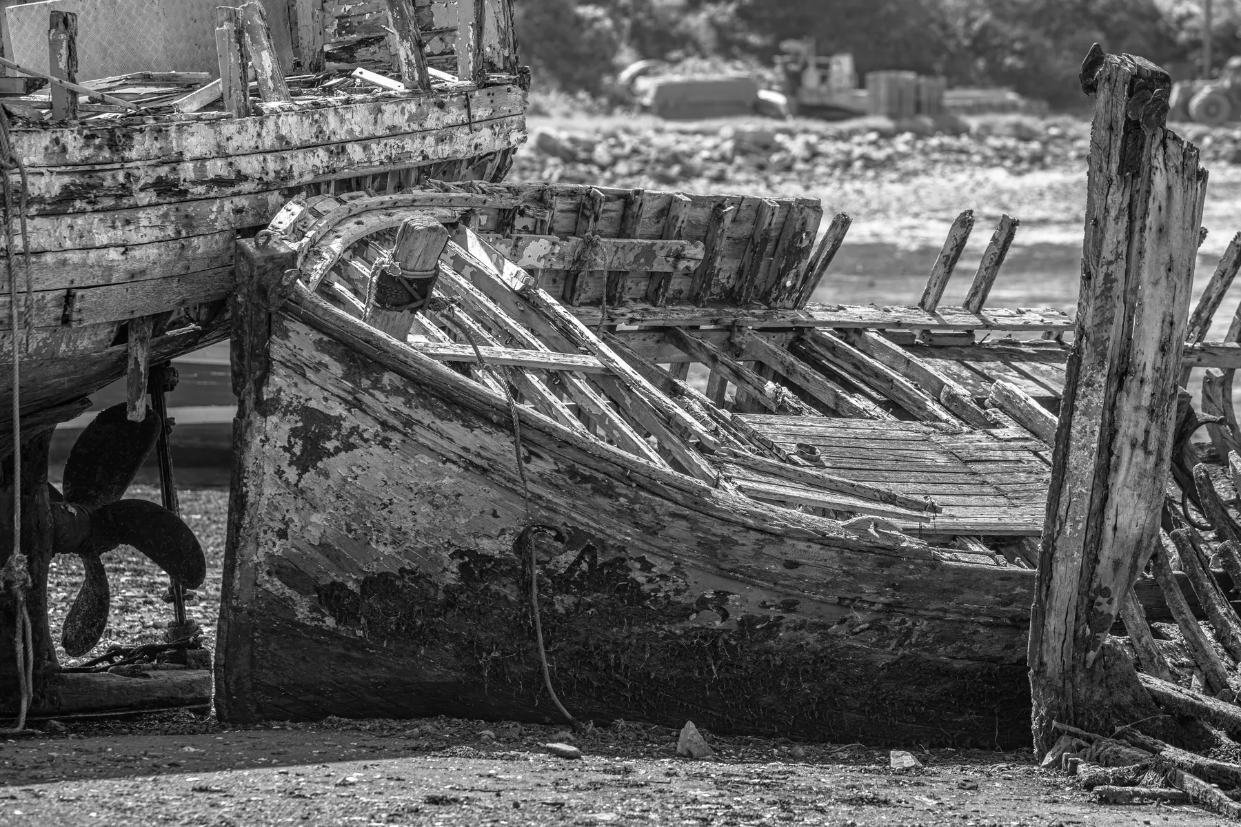 Schwarz-Weiß-Foto eines alten, verwitterten Holzboots mit abblätternder Farbe und freiliegenden Rippen, das an Land liegt und teilweise von anderen verfallenden Holzkonstruktionen umgeben ist.