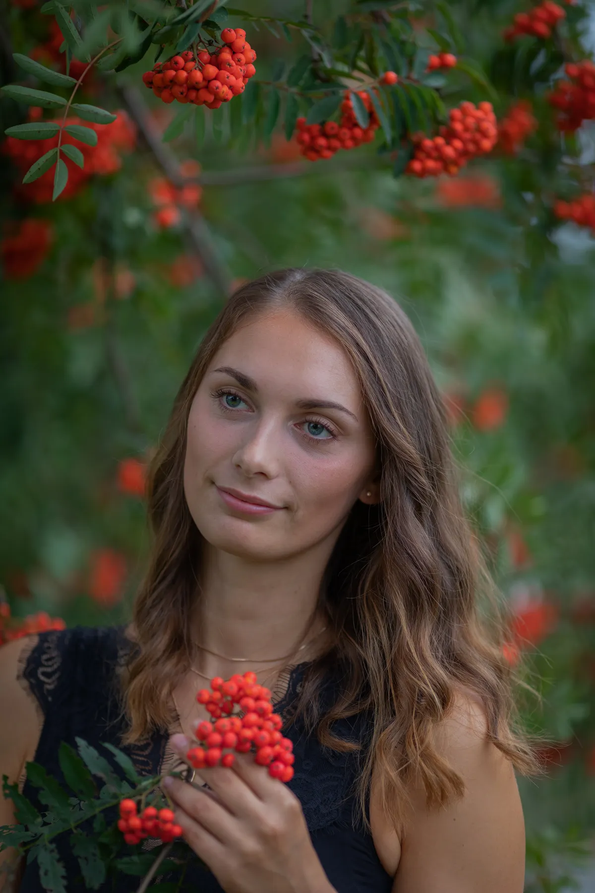 Eine junge Frau mit gewelltem braunem Haar steht im Freien und hält einen Strauß leuchtend orange-roter Vogelbeeren, umgeben von grünen Blättern und weiteren Beerenbüscheln. Sie trägt ein ärmelloses schwarzes Oberteil und schaut sanft in die Kamera.