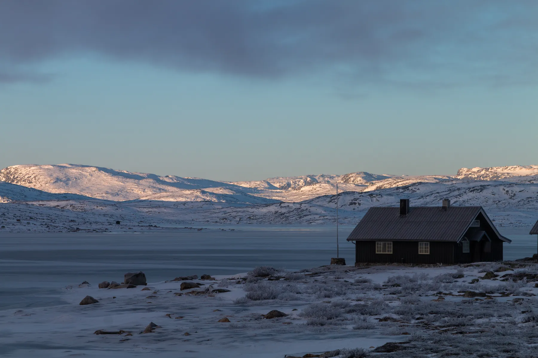 Eine dunkle Holzhütte liegt in der Nähe eines zugefrorenen Sees in der Hardangervidda, umgeben von schneebedecktem Boden und Felsen, mit fernen sonnenbeschienenen Bergen unter einem teilweise bewölkten Himmel.